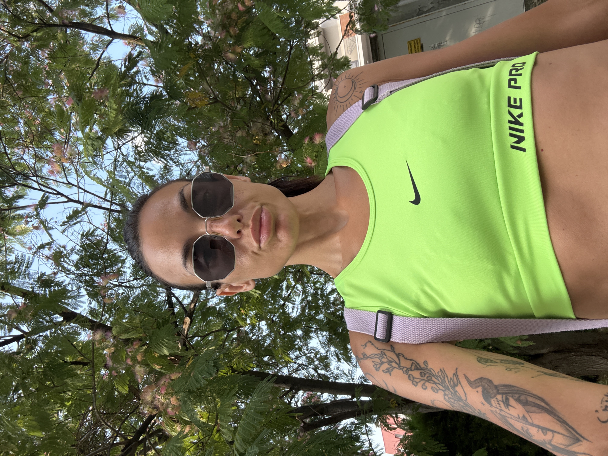A woman with dark hair, wearing sunglasses, and a neon green Nike Pro sports bra stands outdoors. She has tattoos on her arms and shoulders. She is looking towards the camera with a neutral expression. The background consists of lush green foliage and a bright blue sky, suggesting it is daytime and the weather is pleasant. The leaves appear feathery, and some small pink flowers are visible on the branches, indicating it might be late spring or summer. A light purple strap, likely from a backpack or bag, is visible across her shoulder. Some text is visible on the sports bra: "NIKE PRO". The scene appears to be set in a park or garden.