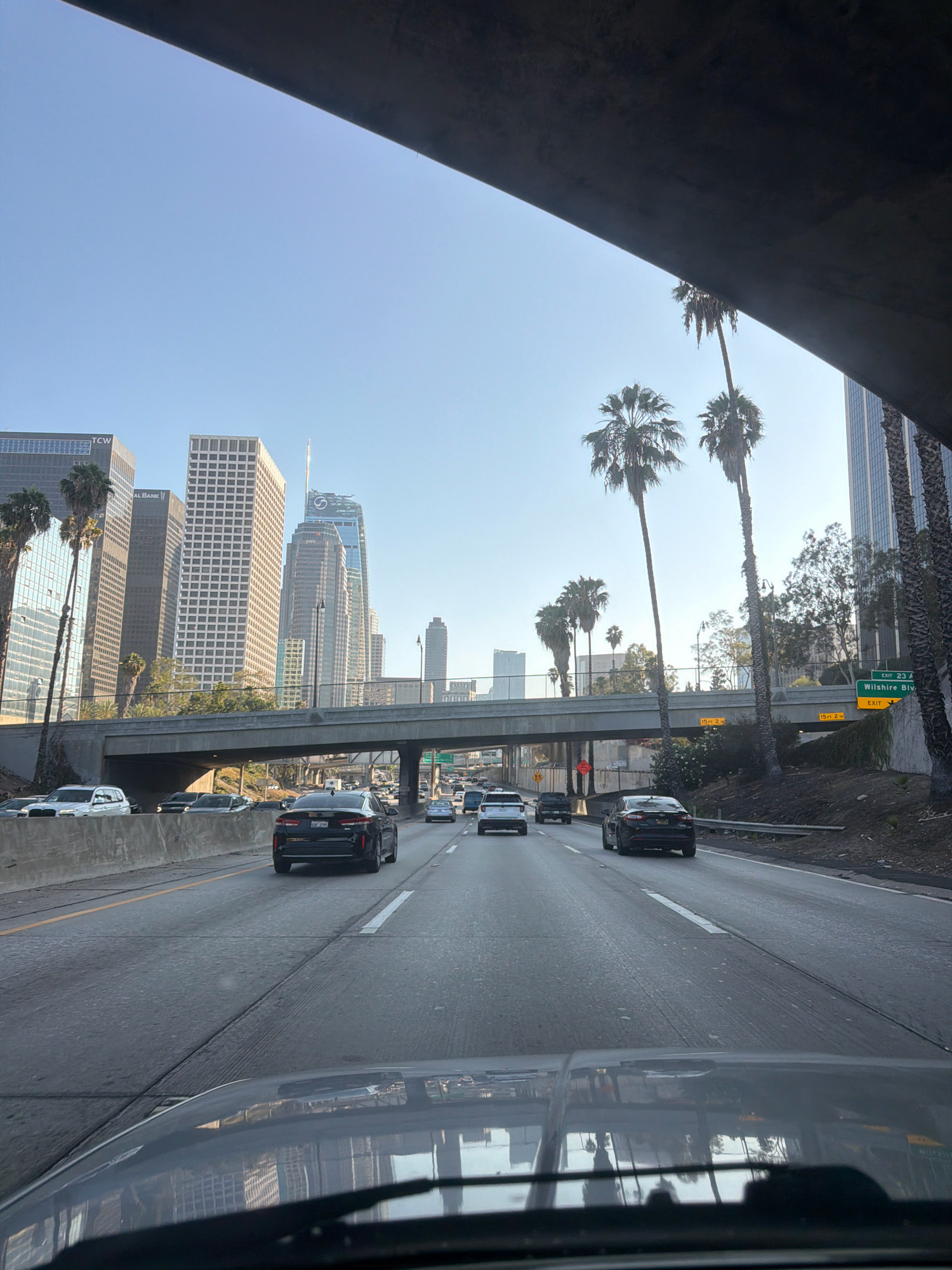 A first-person perspective from inside a vehicle shows a multi-lane highway in Los Angeles, California, United States of America. The highway is congested with traffic, with several cars visible in various lanes. In the background, a cityscape with numerous skyscrapers and palm trees is depicted under a clear blue sky. The vehicle is driving on a freeway, indicated by the presence of overhead signs and multiple lanes of traffic. The weather appears to be sunny and pleasant.