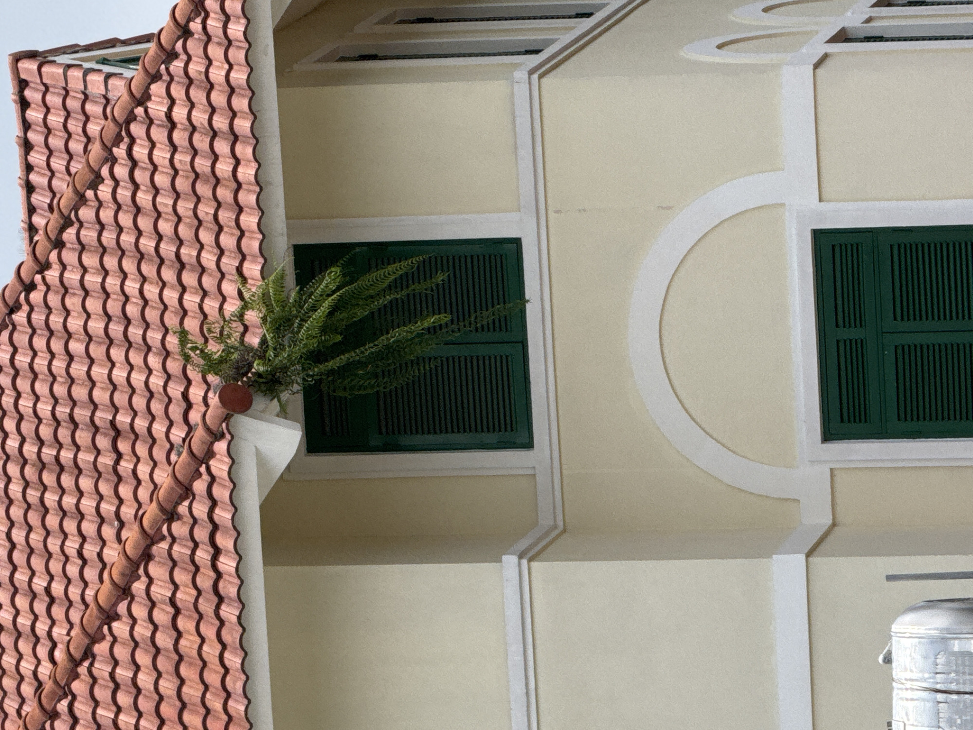 A building in Da Lat, Vietnam features a red tiled roof and light yellow walls. A dark green shuttered window is visible on the upper story, with a hanging fern plant in a brown planter affixed to the eave above it. White decorative molding runs horizontally along the wall, and an arched white molding frames another dark green shuttered window on the lower story. Partially visible in the bottom left corner is a section of a metallic cylindrical tank, possibly a water tank or a fuel tank, with vertical pipes extending upwards. The sky is overcast and light gray.