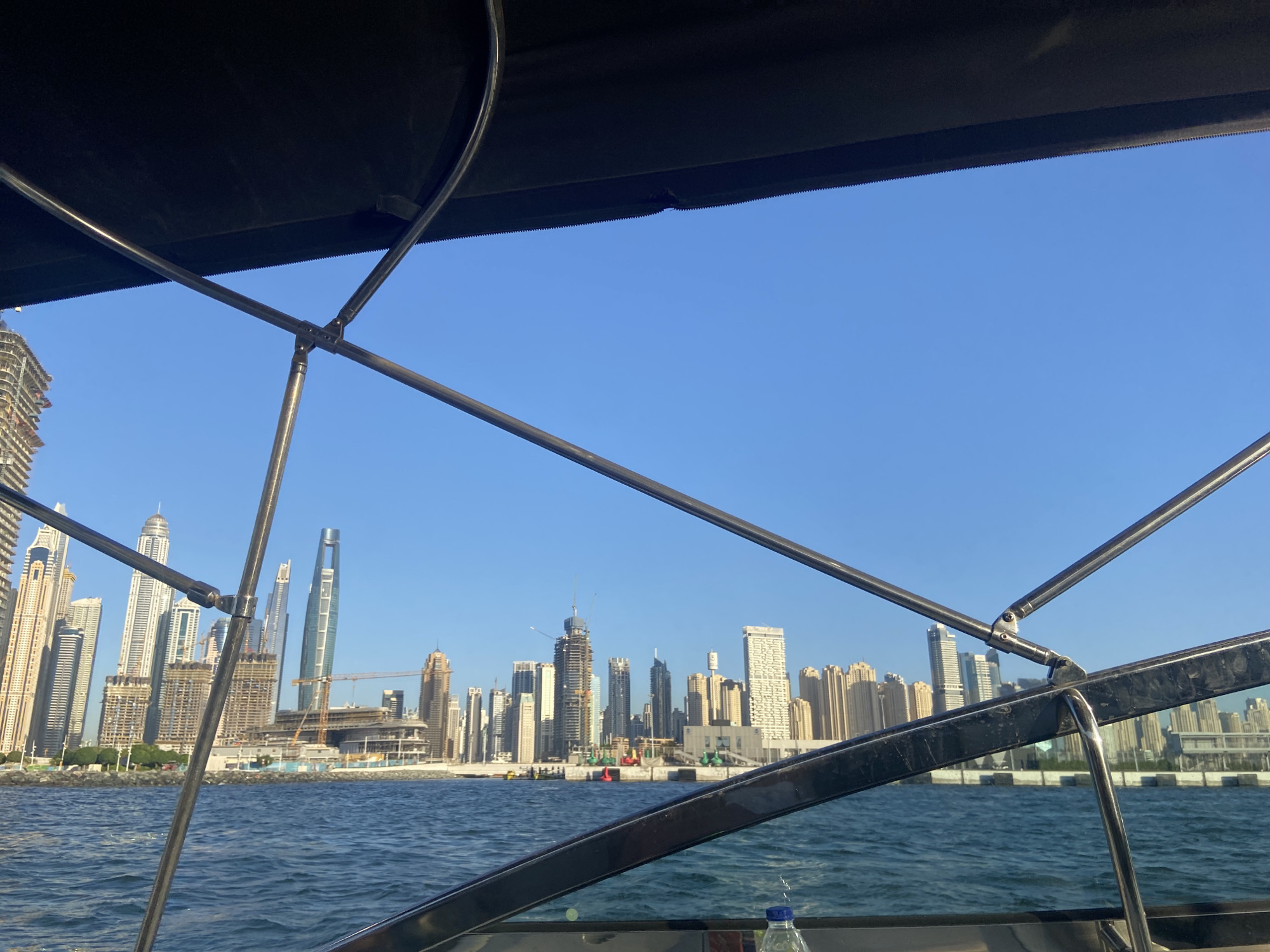 The image depicts a view of the Dubai skyline, including skyscrapers and buildings under construction, as seen from a boat on the water. The foreground includes parts of the boat's frame and structure, suggesting the photo was taken from inside the boat. The sky is clear and blue, and the water is moderately choppy. The buildings line the shore, indicative of an urban coastal area. A bottle is partially visible in the lower right corner of the image, inside the boat.