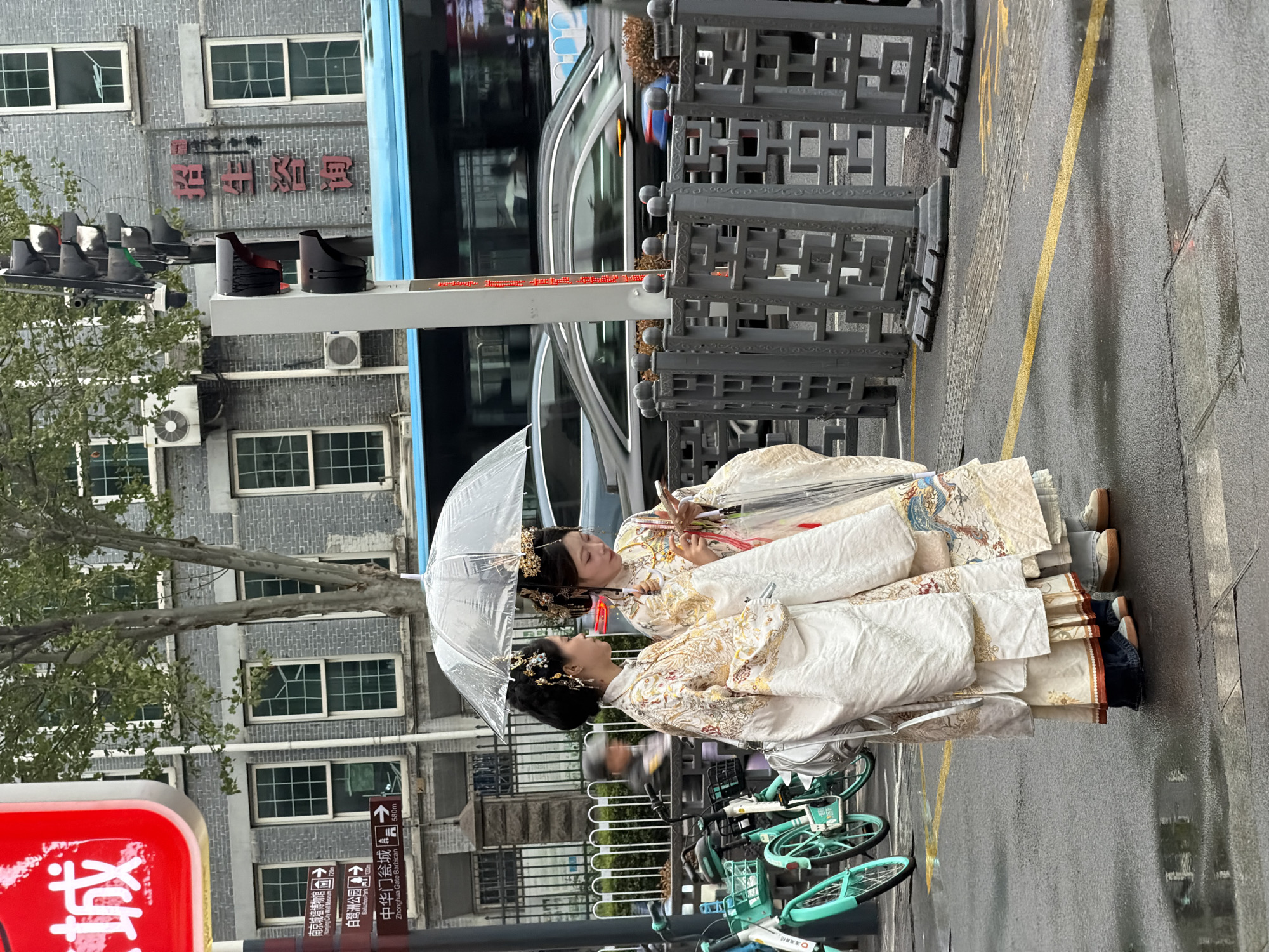 The image shows two people dressed in what appears to be traditional Chinese clothing. They are standing on a street, possibly waiting, under a clear umbrella. The surrounding area is urban, with buildings and a bus visible in the background. Street signs indicate that the location is near Nanjing City Wall Museum, Bailuzhou Park, and Zhonghua Gate Barbican, all located in the Qinhuai District of Nanjing, China. There are also several shared bikes nearby. The street appears wet, suggesting that it has recently rained.