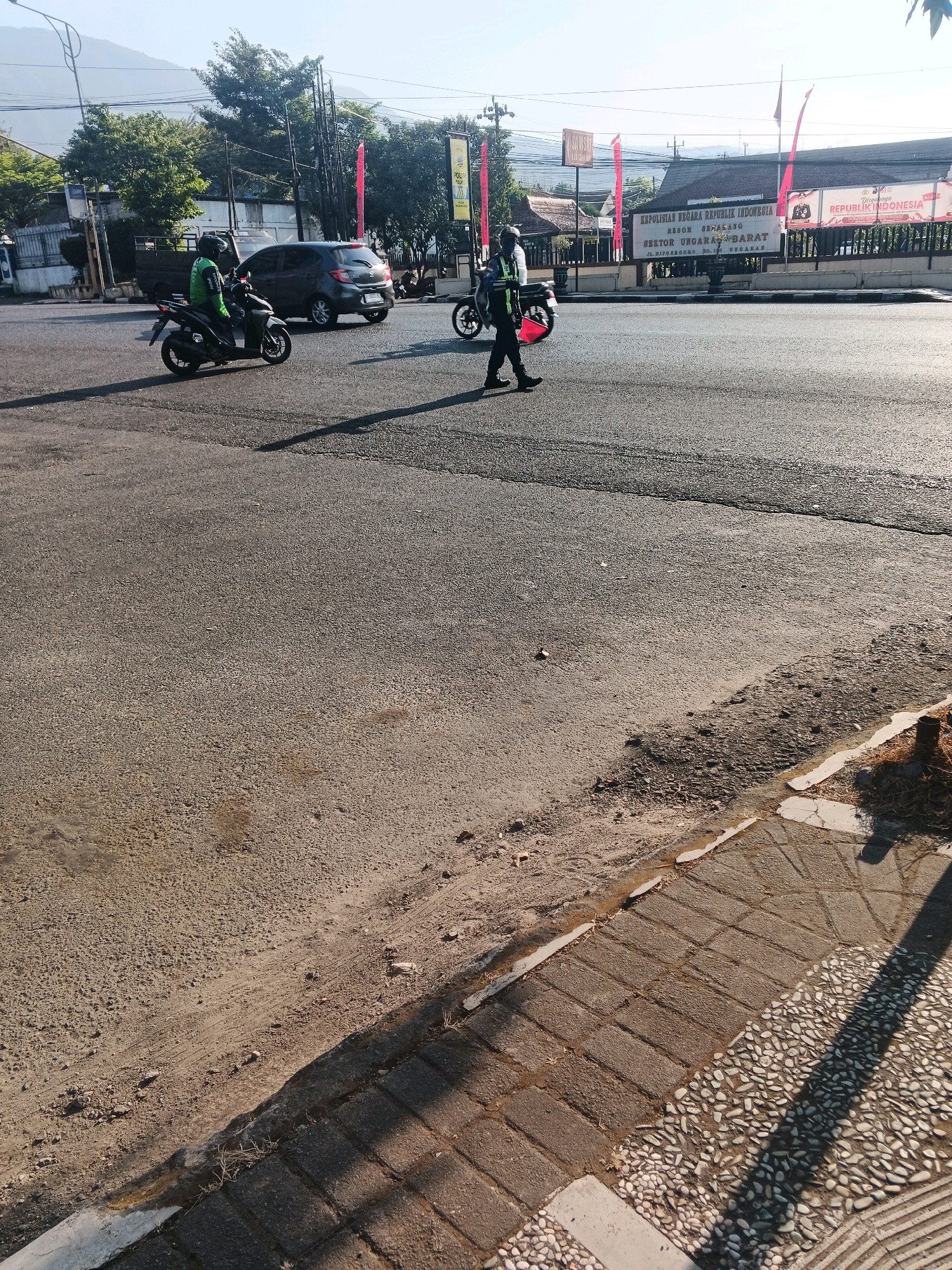 The image depicts a street scene in Ungaran, Indonesia. In the foreground, a paved road with visible tire marks and scattered debris is present. To the right, a cobblestone sidewalk runs alongside the road, bordered by a concrete curb. In the middle ground, a person in a green vest, likely a delivery rider, is operating a motorcycle and crossing the street. Another individual, also wearing a vest and carrying a motorcycle helmet, is walking across the road in the opposite direction. Further in the background, a dark-colored car is parked on the roadside, and a building with signs identifying it as "KEPOLISIAN NEGARA REPUBLIK INDONESIA RESOR SEMARANG SEKTOR UNGARAN BARAT" is visible, indicating this is a police station. Red flags are mounted on poles in front of the building. The environment suggests a sunny day with clear skies, and mountains are faintly visible in the distance.