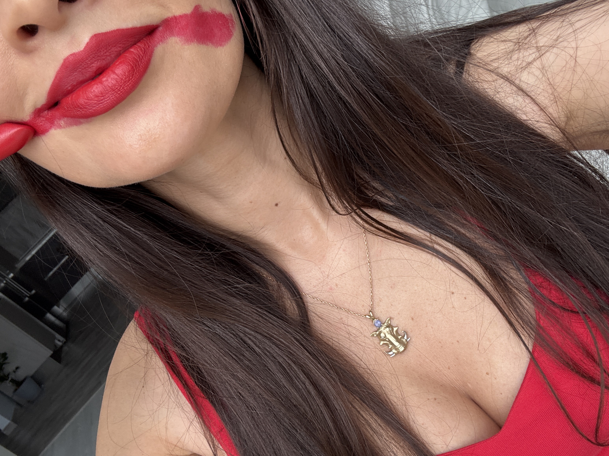 The image is a close-up shot of a person's face and upper body, with a focus on their mouth. Red lipstick is smeared across the lips and cheek, creating a dramatic effect. The person has long, dark brown hair, which falls around their shoulders and chest. They are wearing a red top and a gold necklace with a pendant that appears to be an animal or mythical creature. The background is blurred but suggests an indoor setting with some furniture and possibly a window. The image does not provide any information about the location in the city of Khimki, Russia.