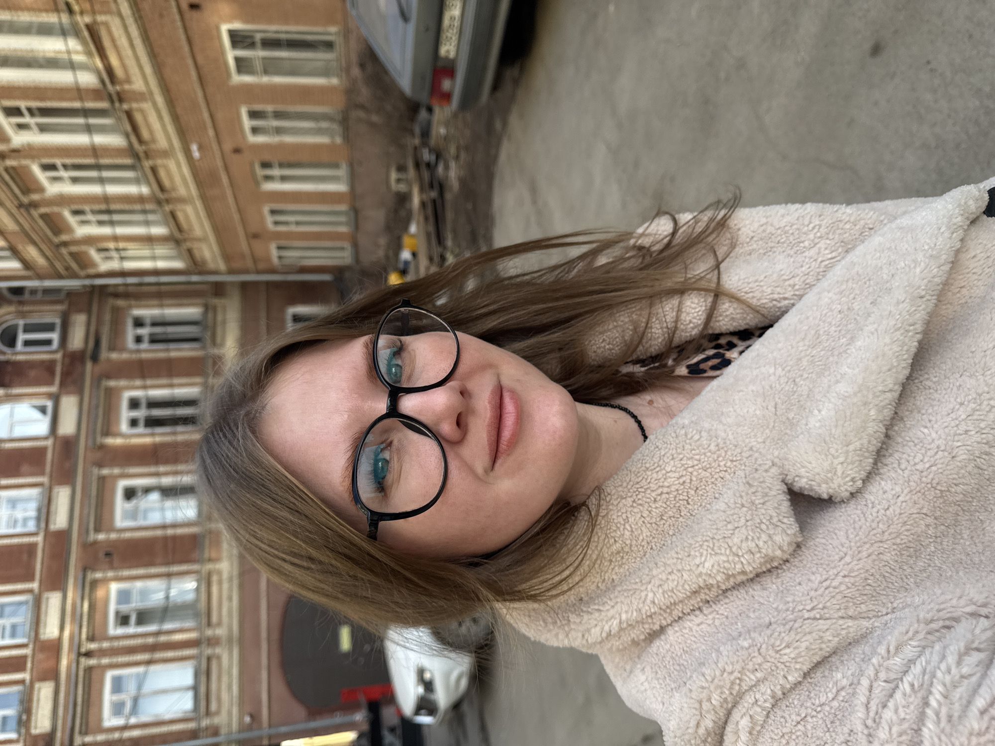 A selfie-style image shows a young woman with light brown hair and fair skin. She is wearing round, black-rimmed glasses. A cream-colored, fuzzy jacket with a collar is draped around her. Underneath, a hint of a leopard-print garment is visible. She's wearing a black beaded necklace.
The background reveals a city street with multi-story brick buildings. These buildings feature multiple rows of windows. In the upper part of the frame, a portion of a parked vehicle is visible, partially obscured. The scene is possibly taken in Perm, Russia. The light suggests it is daytime.