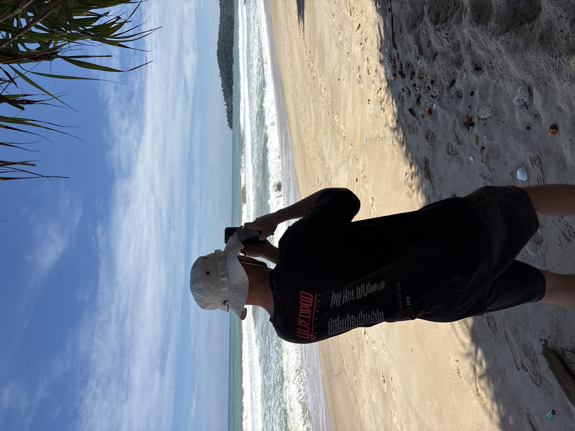 A person wearing a white bucket hat and a black t-shirt is standing on a sandy beach in Choeng Thale, Thailand, taking a photo with a smartphone. The person is facing the ocean, which has waves and a distant coastline with trees. The sky is blue with scattered white clouds. The sand is light brown with some debris, including plastic cups and orange pieces. Shadows from palm fronds are cast on the sand. The black t-shirt has red text and graphics on the back, including the words "YUEDPOOD" and "PARADISE ON TOUR 2023".