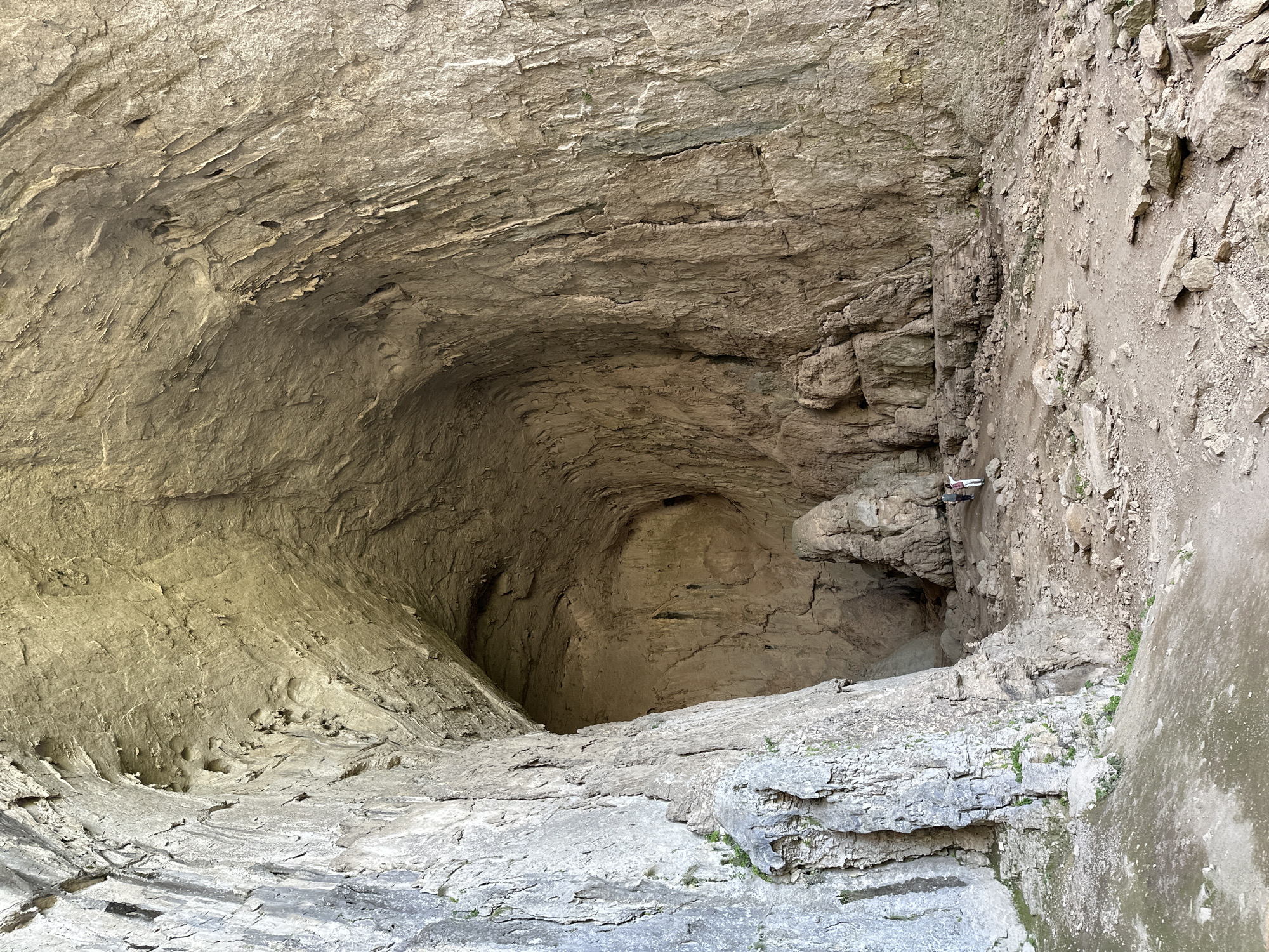 This is a photograph of a cave or crevice located in Karlukovo, Bulgaria. The area is surrounded by rugged, light-brown rock formations. There is a smaller cave entrance visible within the larger cavern. A white and black object, possibly a drone, is visible near the entrance of the smaller cave. The ground is also rocky, composed of various sizes of grey and light-brown stone.