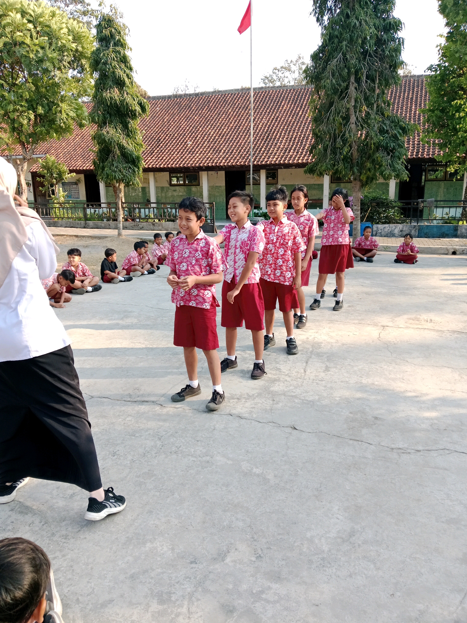 In Salatiga, Indonesia, at what appears to be a schoolyard, a group of children in uniform are gathered outdoors. Several boys are standing in a line, wearing matching red shirts with a white floral pattern and red shorts. They are facing to the left of the frame, with some smiling and looking towards a person out of frame. Behind them, other children are sitting on the ground, also in uniform. A woman in a white shirt and black skirt is visible in the foreground, facing away from the camera and towards the children. In the background, a school building with a terracotta tiled roof is situated behind a fence and some trees. A red flag flies on a flagpole to the left of the building. The ground is paved with concrete.