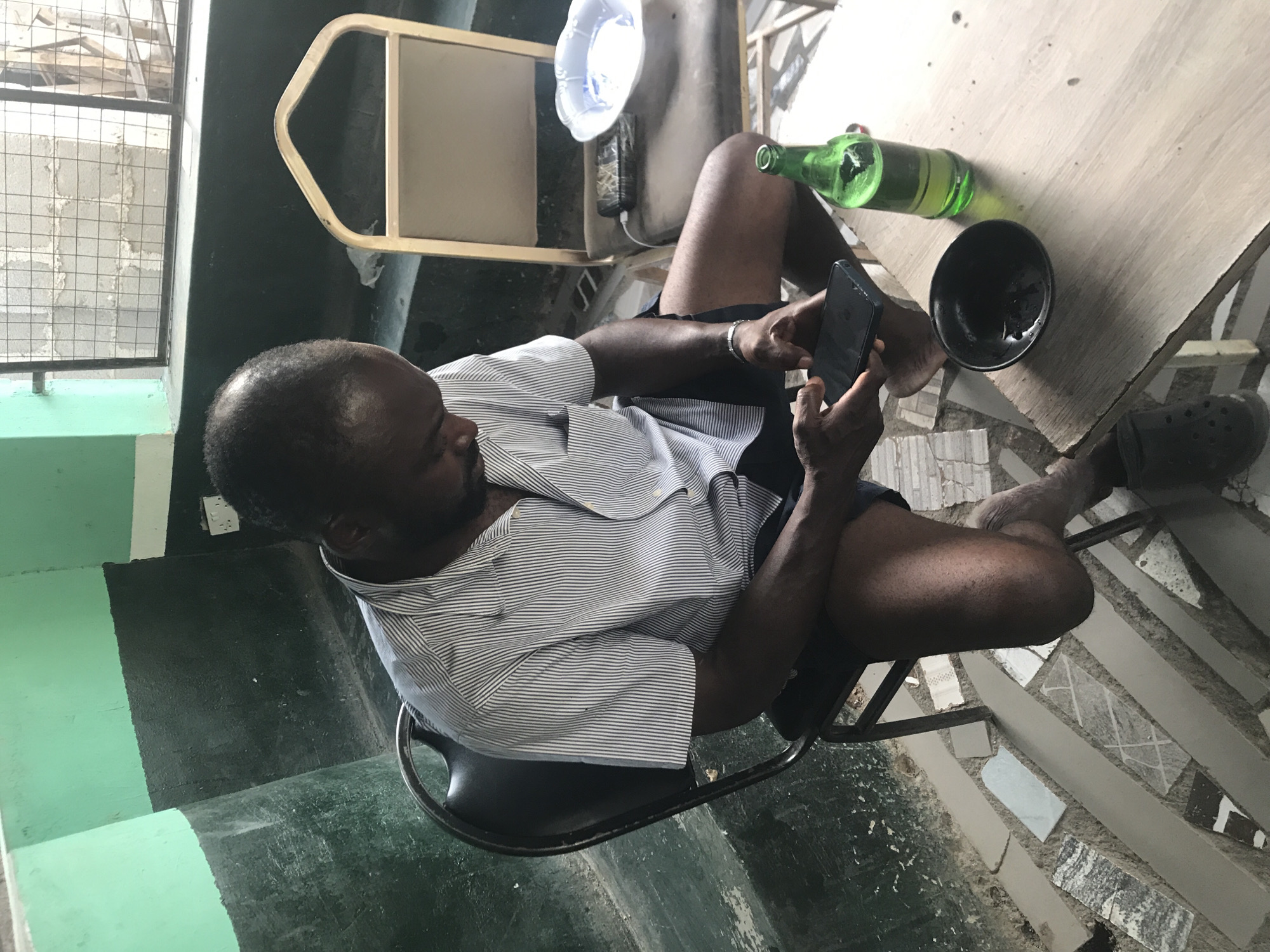 An adult Black man is seated on a black metal chair in what appears to be an outdoor or semi-outdoor setting. He is facing a table and holding a black mobile phone, which he is looking at. He is wearing a short-sleeved, light-colored striped shirt and dark shorts. Next to him on the table is a green glass bottle lying on its side, and a black bowl. The table is light-colored and appears to be made of wood. A single black Croc shoe is near the table leg. The flooring consists of broken tiles. The location may be in the city of New Stadium, Nigeria.