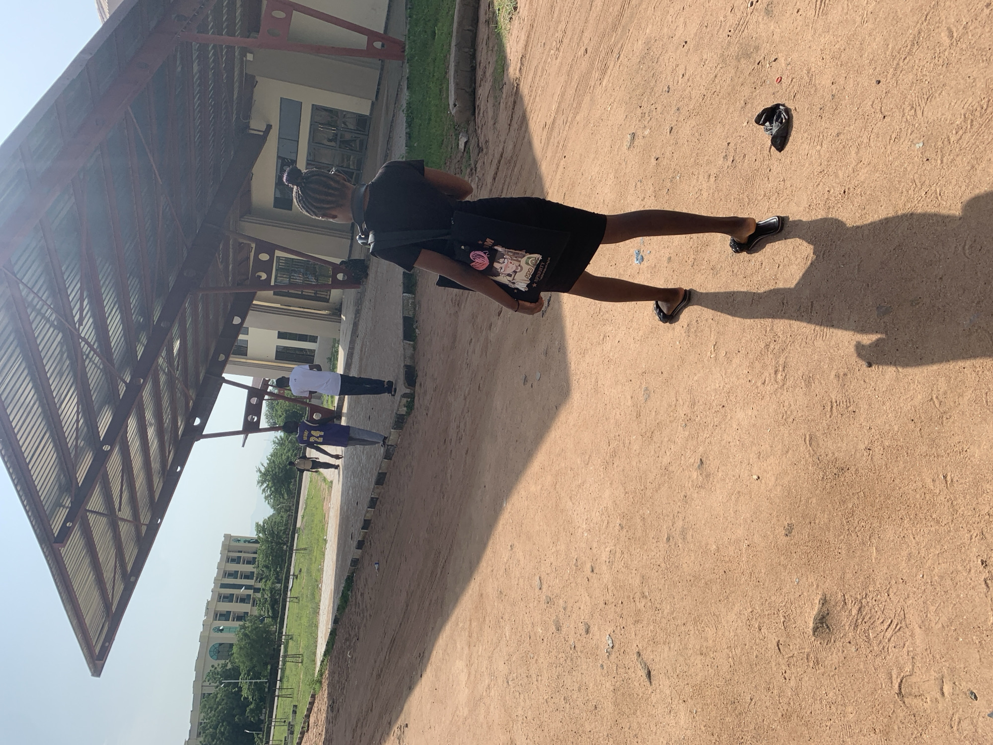 In the city of Jalingo, Nigeria, a young Black female is captured from behind, walking away from the camera on a dirt path. She is wearing a black dress and black sandals, and carrying a black tote bag with cartoon imagery. Her hair is styled in braids and pulled into a bun. She is walking towards a covered walkway with a corrugated metal roof and a red-brown metal support structure. In the background, two other individuals are walking away from the camera, with one wearing a purple jersey with the number 24. Behind them, a multi-story building with a beige facade and numerous windows stands amidst a green, grassy landscape. The overall environment appears to be an outdoor area with sparse vegetation and a clear, bright sky, suggesting a warm, sunny day.