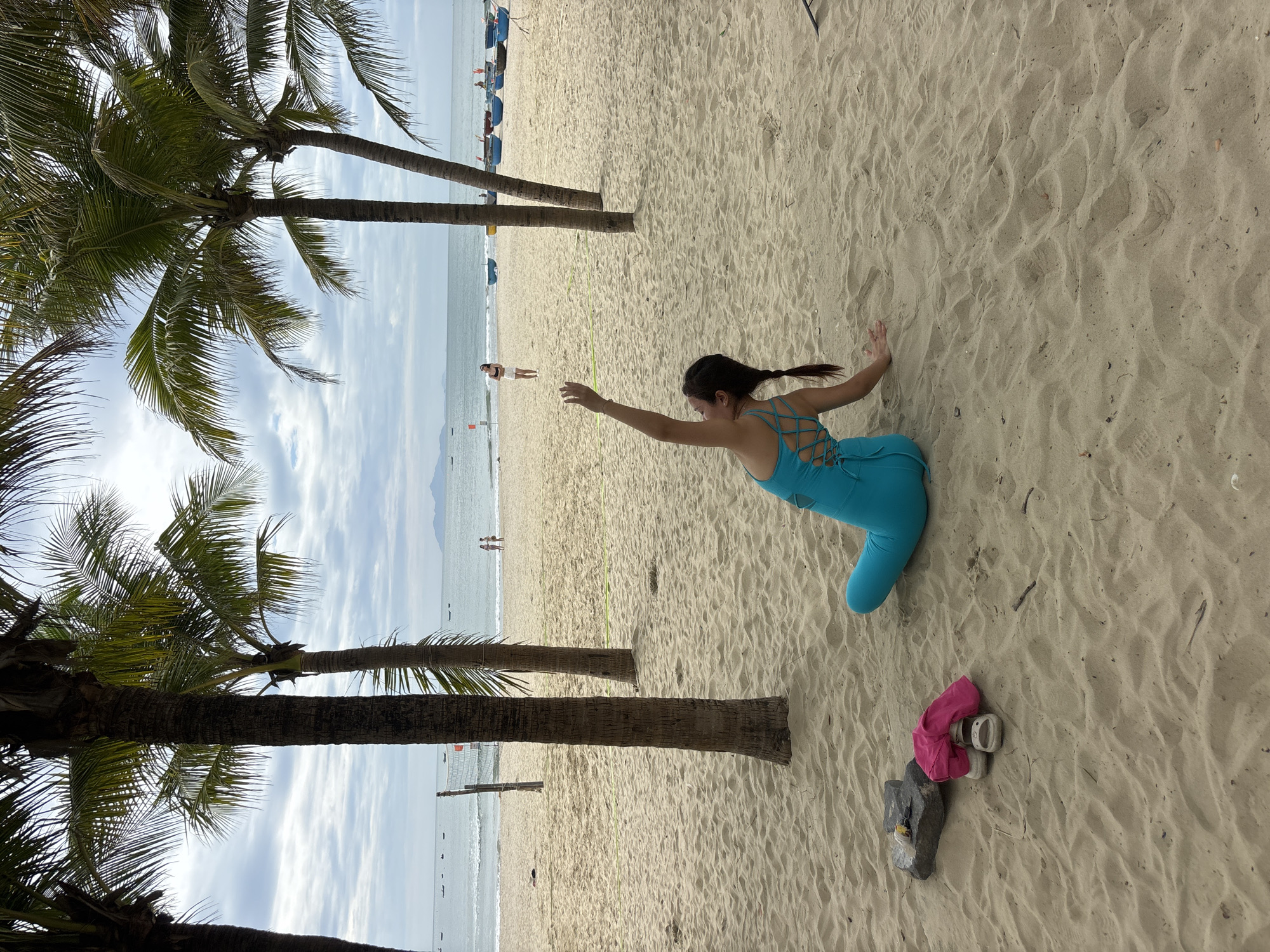 The image shows a woman practicing yoga on a sandy beach with palm trees. In the background, the ocean is visible with several boats and a distant island. There are other people in the background, some walking along the shore and others near the boats. The woman is wearing a turquoise athletic outfit and is in a seated pose with her arms extended. The beach appears to be in a tropical or subtropical location, likely a tourist area given the presence of boats and the general atmosphere. Based on the palm trees, ocean, and boats, this could be a beach in Đà Nẵng, Vietnam, such as My Khe Beach, known for its long sandy stretches and palm-lined shores.