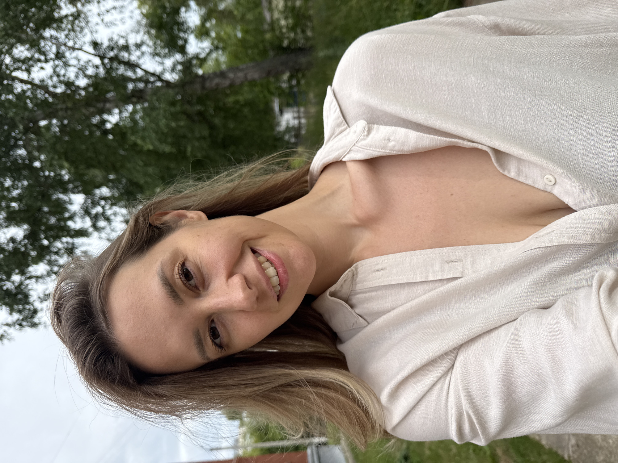 The image shows a close-up, angled shot of a woman with blonde hair and fair skin. She is smiling broadly, her head tilted to the side. She is wearing a light-colored, possibly linen, button-up shirt that is partially unbuttoned, revealing her collarbone and upper chest. The background is blurred, showing green foliage and a cloudy sky, suggesting an outdoor setting, likely in a park or garden. The lighting appears to be natural, indicating it is daytime. There is no visible text in the image. The overall mood of the image is cheerful and relaxed. Given the location context of Chaikovsky, Russia, the scene likely depicts a person enjoying a casual moment outdoors.