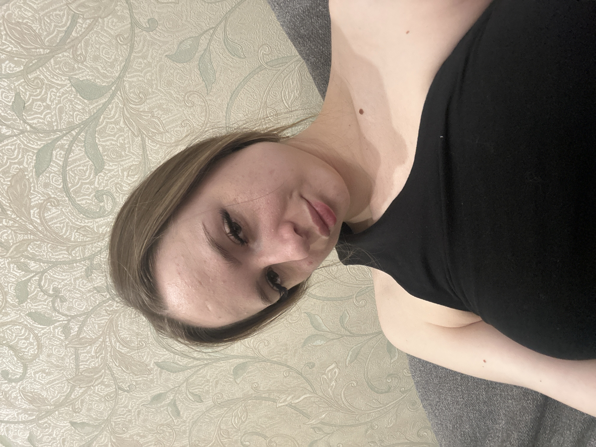 A woman with fair skin and brown hair lies on a patterned surface, wearing a black top. Her head is tilted slightly towards the camera, and she is looking downward. The background consists of a light-colored surface with an intricate, swirling pattern of leaves and vines. Part of a gray surface is visible on the bottom right. The location and city are unknown.