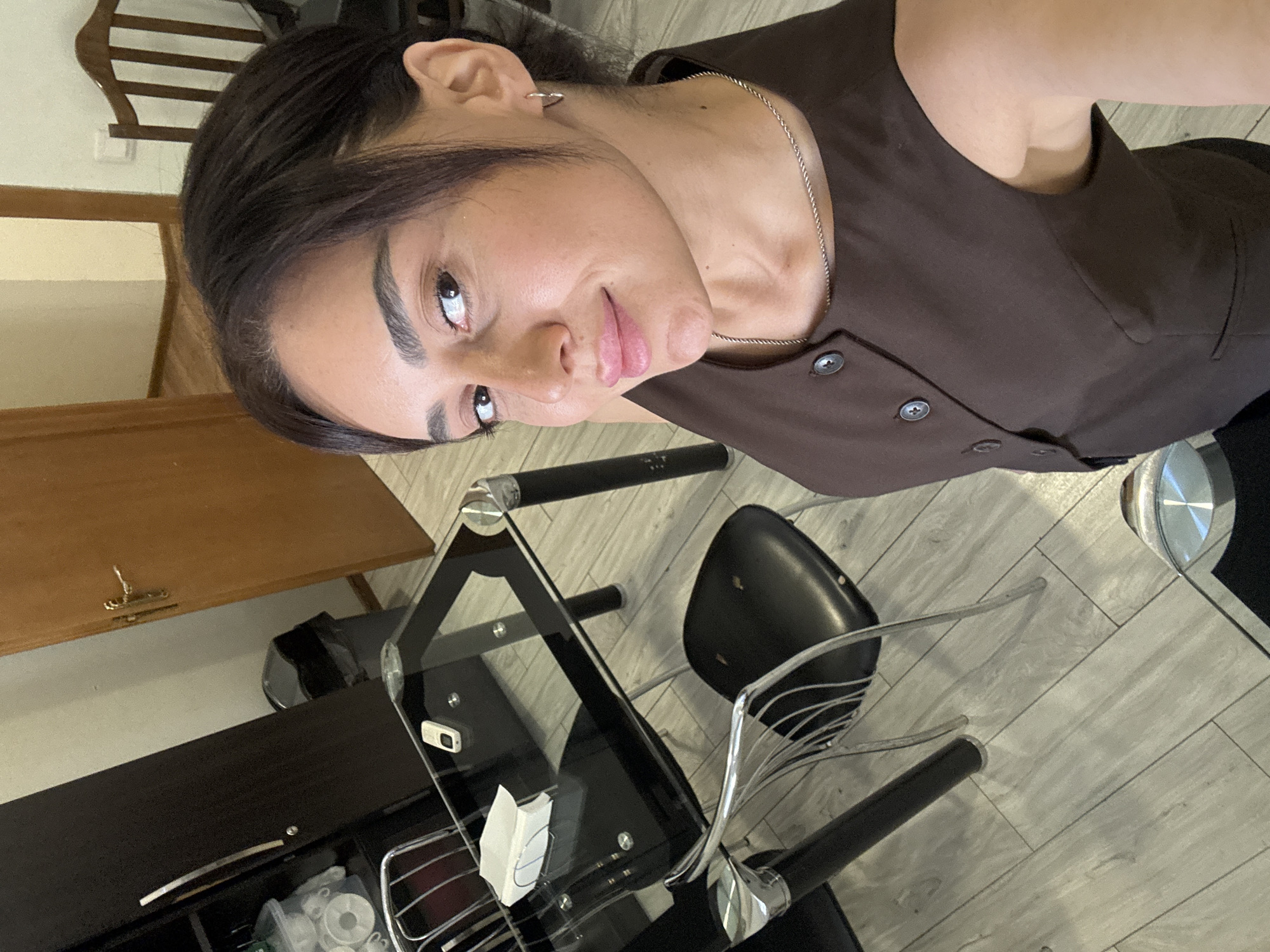 A woman with dark hair and brown eyes is taking a selfie in what appears to be a dining area. She is wearing a brown vest over a white top, and a silver necklace. She is smiling at the camera. In the background, there is a glass-topped dining table with chairs, a dark brown cabinet, a wooden door, and a trash can. The floor is covered with light-colored laminate flooring. The setting appears to be an indoor residential or small commercial space. There is no indication that this location is in the city of Tbilisi, Georgia.