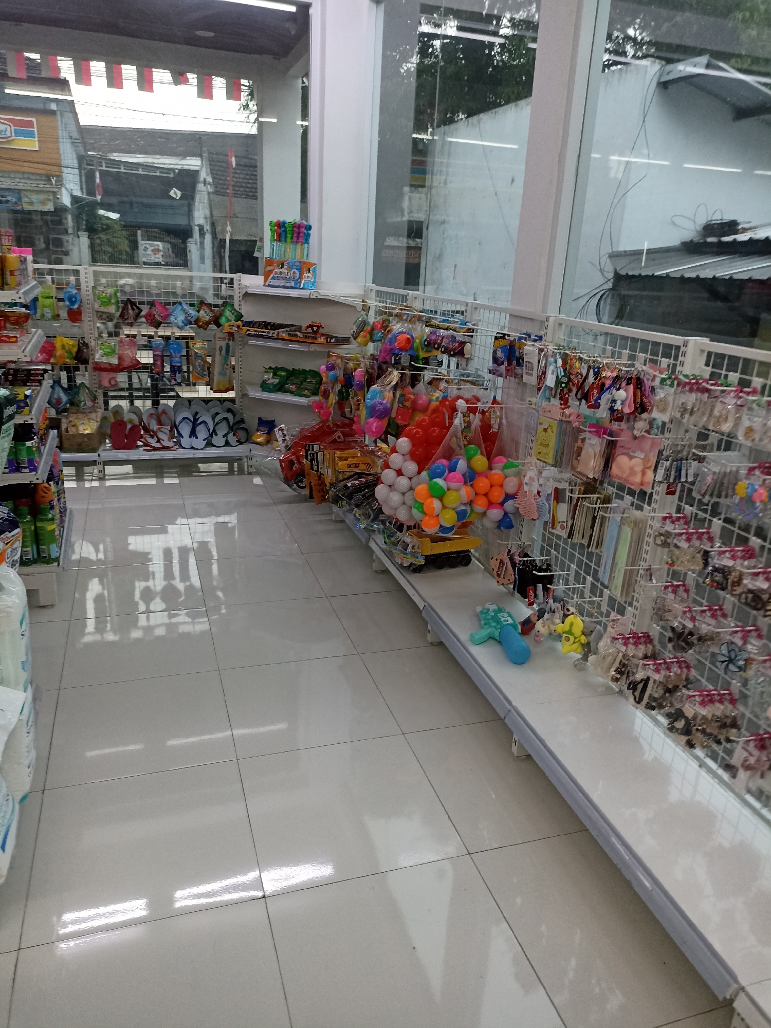 The image depicts the interior of a small convenience store or minimarket, likely in Tompokersari, Indonesia, given the context. The scene is dominated by shelves filled with various merchandise, primarily toys and accessories. On the left side, there are shelves with bottled drinks and other packaged goods. In the center, there is a display of colorful flip-flops and various children's toys, including small trucks and balls. The right side of the store features racks of accessories such as hair clips, keychains, and small trinkets, all neatly packaged. The floor is tiled with large, pale gray tiles that reflect the overhead lighting, creating a bright and clean appearance. The overall impression is that of a well-organized retail space offering a range of affordable items. There are no people visible in the image. The store appears to be open for business, with goods displayed for sale. The exterior of buildings and trees are visible through the large windows, suggesting it is daytime.
