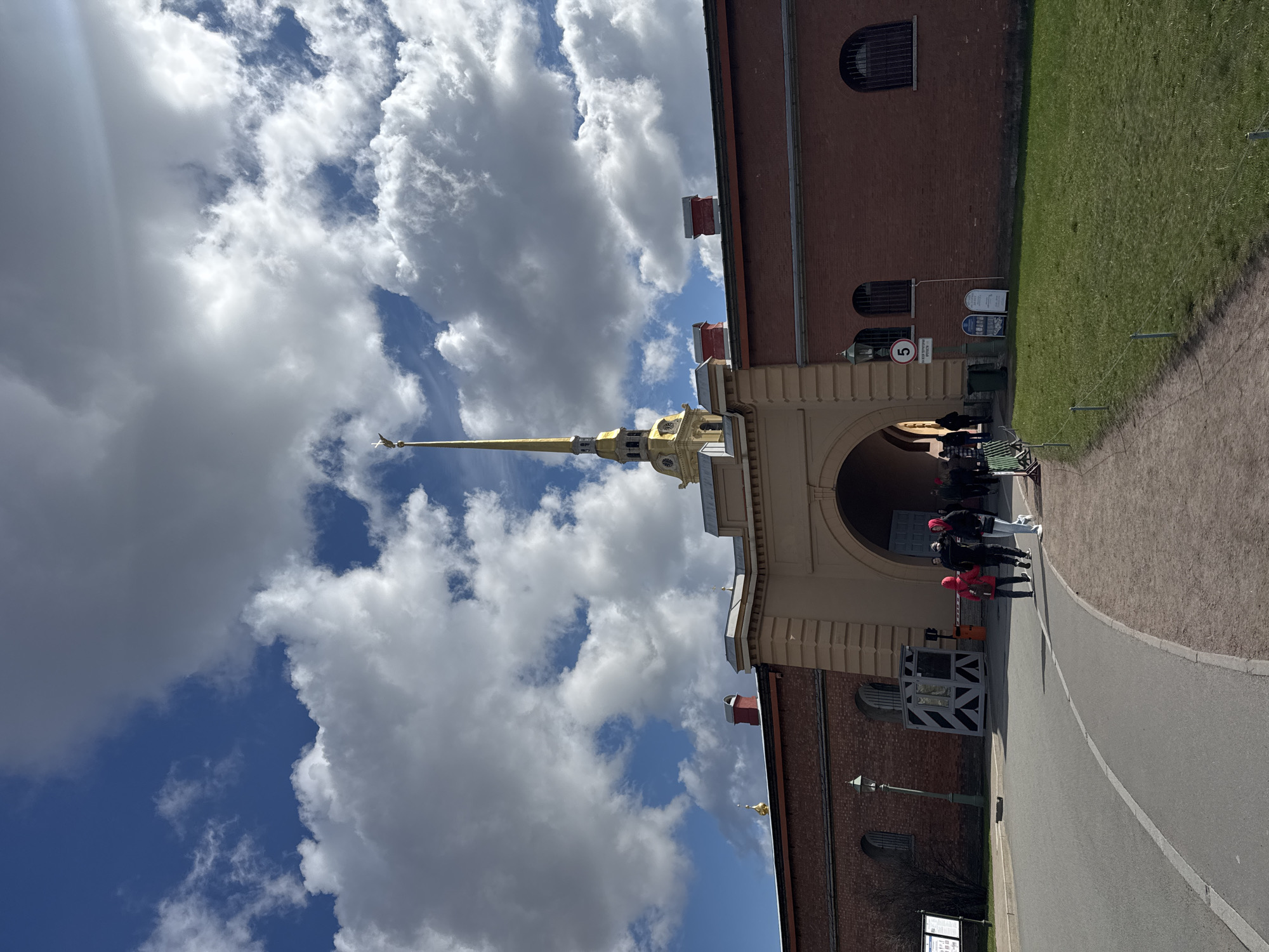 The image depicts the Peter and Paul Fortress in Saint Petersburg, Russia. A golden spire rises against a backdrop of blue sky and white clouds. People are entering the fortress through an archway in a brick building. A guard booth with black and white stripes is visible near the entrance. A paved road leads into the fortress. The sky is partly cloudy.