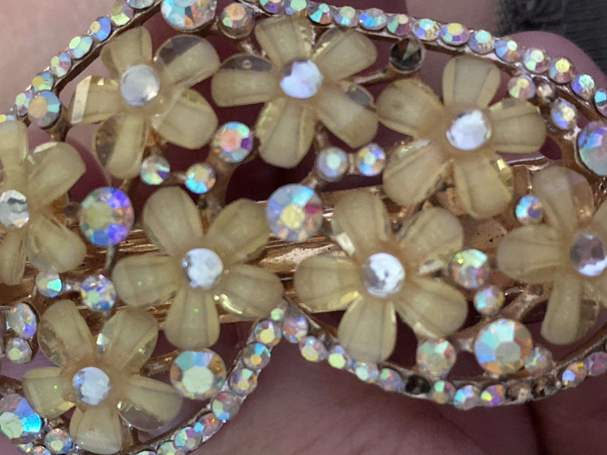 The image presents a close-up, highly detailed view of an ornate accessory, likely a hair clip, barrette, or brooch. The item features a delicate floral design composed of numerous small, translucent light yellow or cream-colored flowers. Each flower is centered with a prominent, round, faceted rhinestone that displays an iridescent "Aurora Borealis" effect, reflecting a spectrum of colors including blue, pink, and yellow. Surrounding these flowers and integrated into the overall structure are many smaller, similarly iridescent rhinestones, all set into a gold-toned metal base. The material of the flowers appears to be a soft, matte plastic or resin. The background, visible in parts, appears to be a person's skin, suggesting the accessory is being held or displayed. No specific activity or event is depicted, nor are there any visible texts. The image focuses entirely on the intricate craftsmanship and sparkling details of the decorative item, with no cues for time of day, weather, or specific location within Novokuznetsk, Russia.