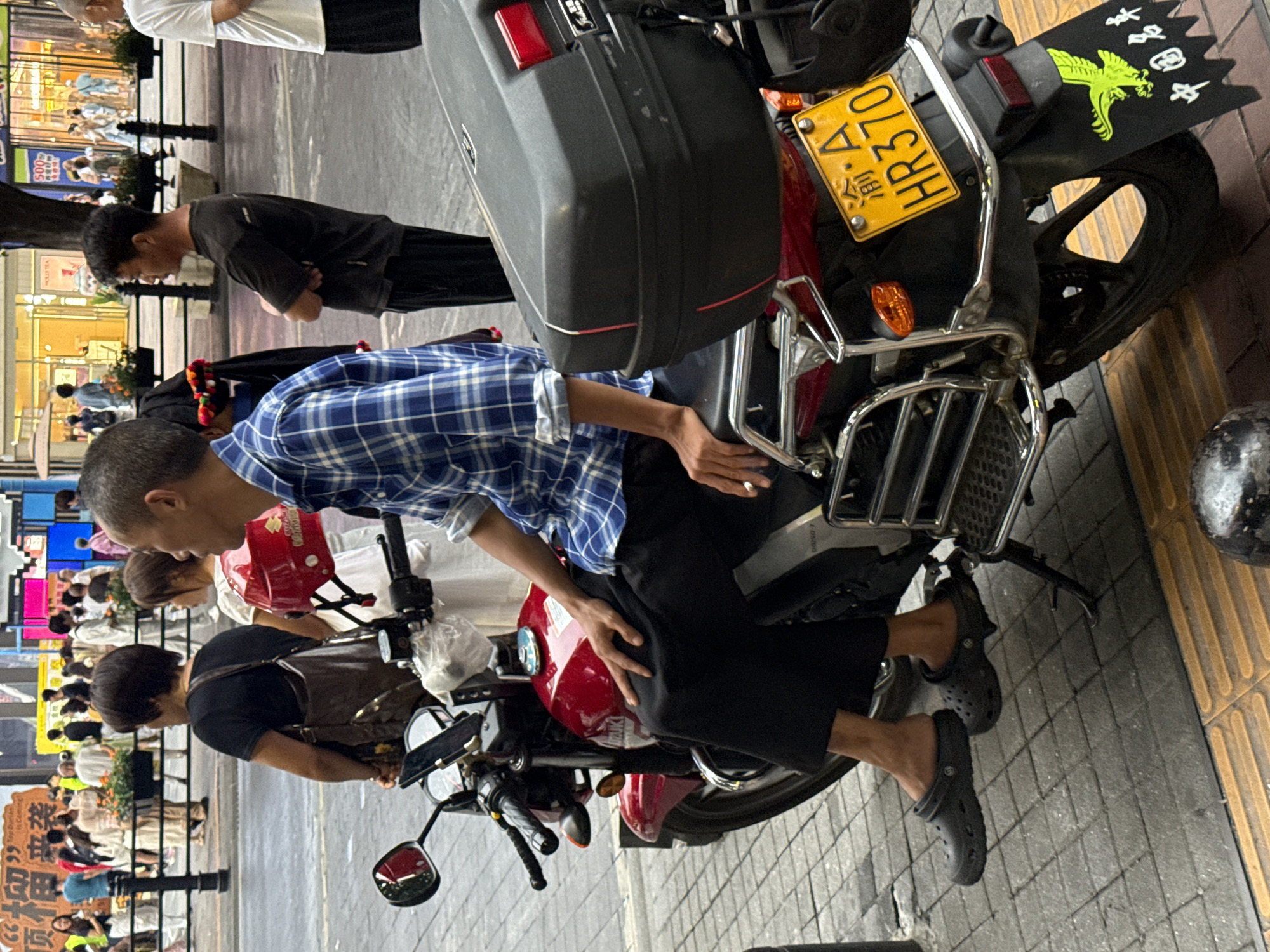 An outdoor, eye-level shot captures a scene on a city street in Yuzhong District, China. In the foreground, a man is seated on a red motorcycle. He is wearing a blue and white checkered shirt and black pants. He is holding a lit cigarette in his right hand, which is resting on his thigh. His feet are shod in black Crocs. The motorcycle has a large black storage box mounted on the rear and a yellow license plate with the characters "渝 A HR370". Behind the man and the motorcycle, a woman in a black shirt and a brown bag slung over her shoulder is visible. Further in the background, a crowd of people are walking on the sidewalk in front of storefronts. The environment is a typical urban street scene with buildings, sidewalks, and traffic.