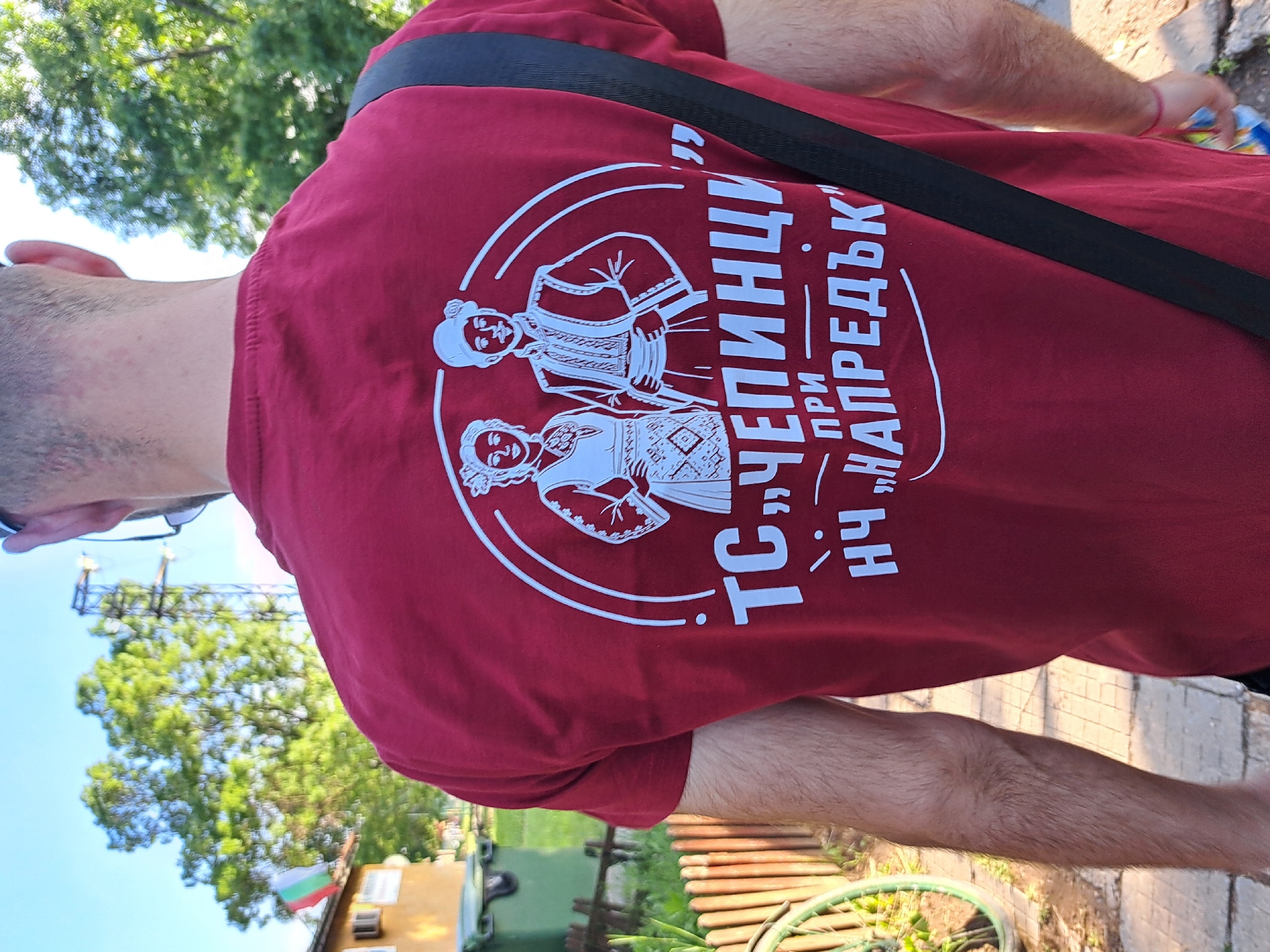 A man is wearing a maroon t-shirt with a design on the back. The design features two figures in traditional Bulgarian clothing within a white circle. Text in Bulgarian surrounds the figures. A black strap crosses diagonally across the man's chest. The man is standing outdoors on a paved surface. In the background, there are trees, a building with a Bulgarian flag, and a structure resembling a light tower. A portion of a green bicycle wheel is visible near the bottom of the frame. The location is likely Negovan, Bulgaria, based on the Bulgarian flag and the t-shirt design.