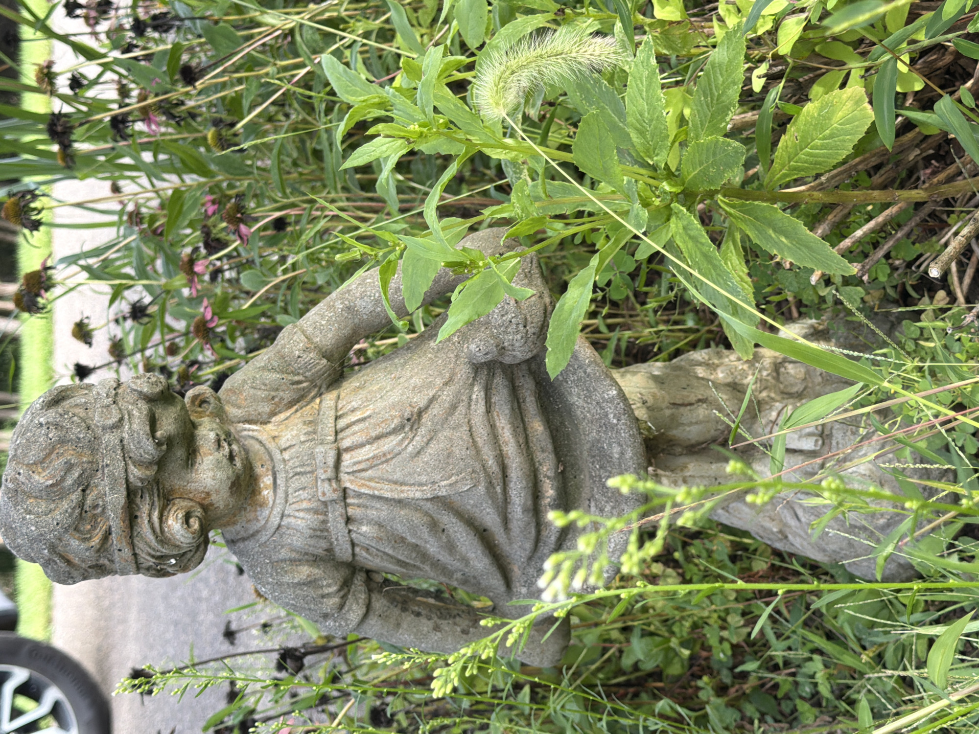 A weathered stone statue of a young child, resembling a girl with curls and wearing a dress, stands amidst overgrown greenery in Middlesex Township, USA. The statue is partially obscured by tall grasses and plants, some of which bear faded purple flowers. To the left, a portion of a tire, likely from a vehicle, is visible on a paved surface. The statue appears to be made of concrete or similar material and shows signs of age and exposure to the elements. The surrounding environment is a naturalistic setting with abundant vegetation, suggesting a garden or unkempt outdoor area. The lighting indicates it is daytime, with sunlight illuminating the scene.