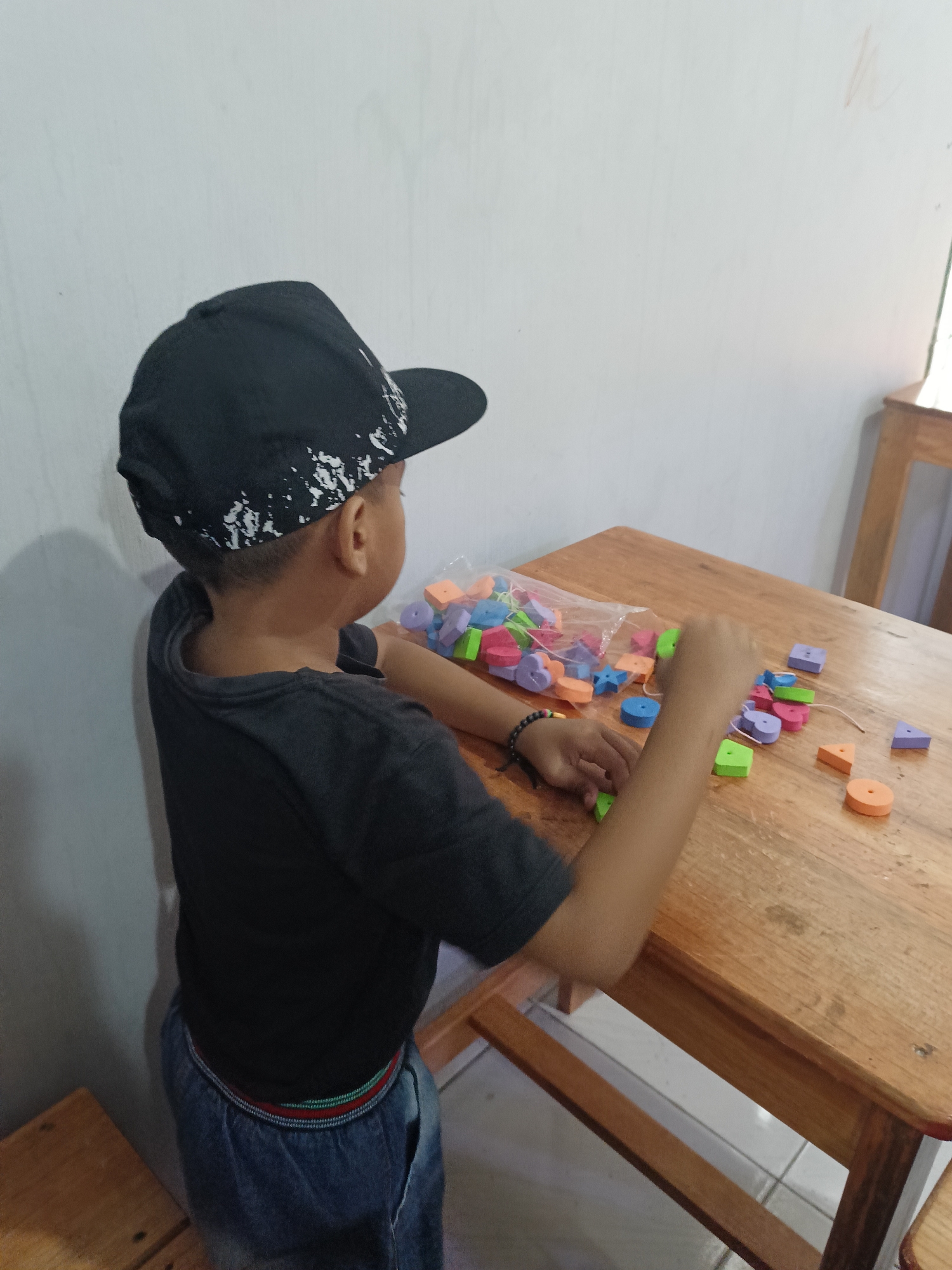 A young boy wearing a black baseball cap and a black t-shirt is playing with colorful foam letter and shape toys on a wooden table. The boy is on the left side of the image, his back to the viewer. He is reaching out with his right hand towards the toys. The toys are scattered on the table, some in a clear plastic bag. The table is made of light-colored wood. The background is a white wall. The scene appears to be indoors, likely in a home or educational setting. The overall setting is consistent with a scene in Palangka Raya, Indonesia.