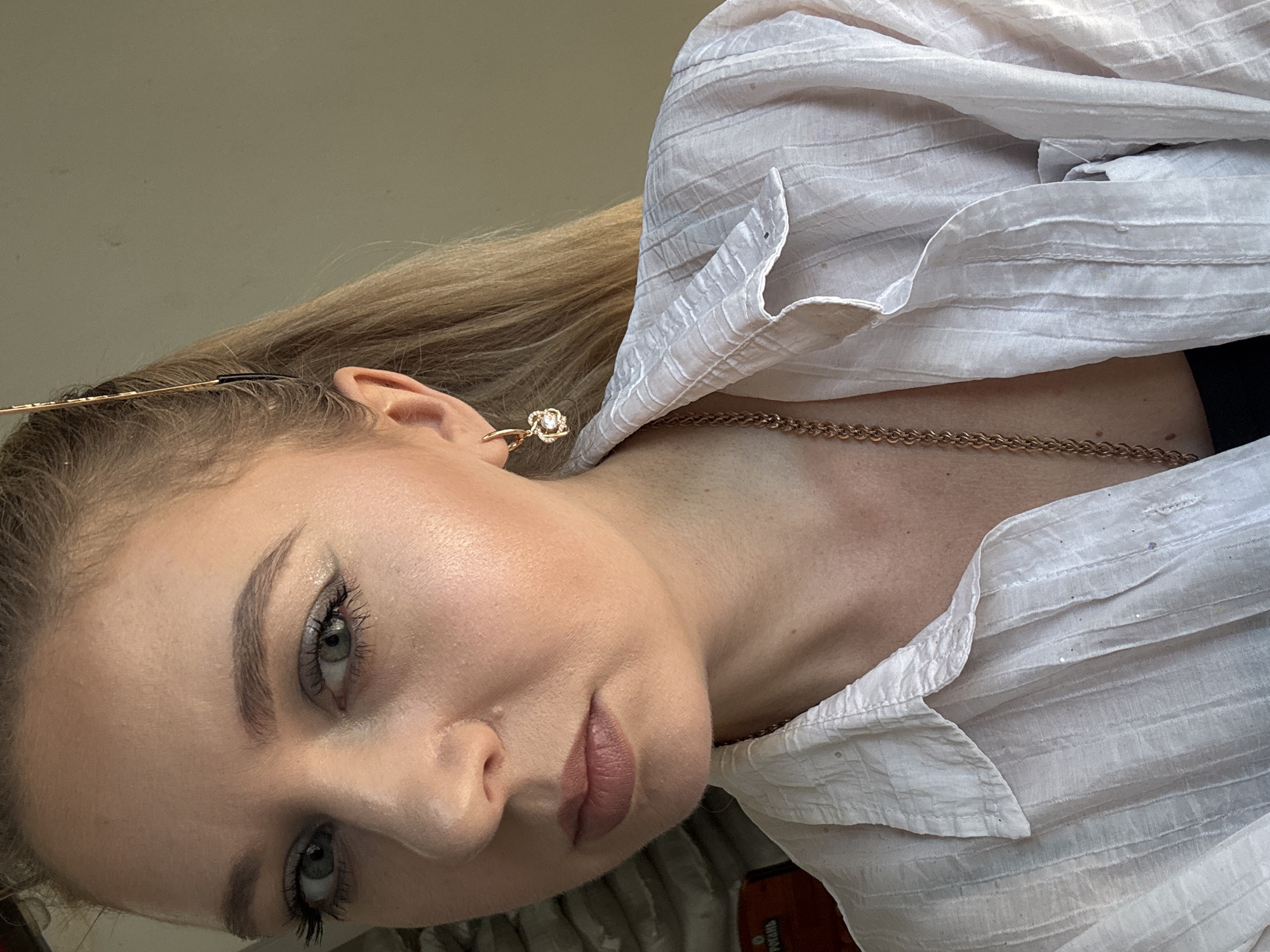 A close-up, medium shot captures a young Caucasian woman from the chest up, looking directly at the camera. She has fair skin, light blue eyes with visible makeup, and light brown hair pulled back with a gold barrette. Her mouth is closed, and she wears a light pink lipstick. A gold hoop earring with a pearl and small diamonds adorns her left ear. She is wearing a white, textured button-up shirt and a gold chain necklace. The background is a neutral, light brown wall. The lighting appears to be natural, coming from the side, casting soft shadows on her face. The setting is indoors, and there are no discernible city or environmental details present.