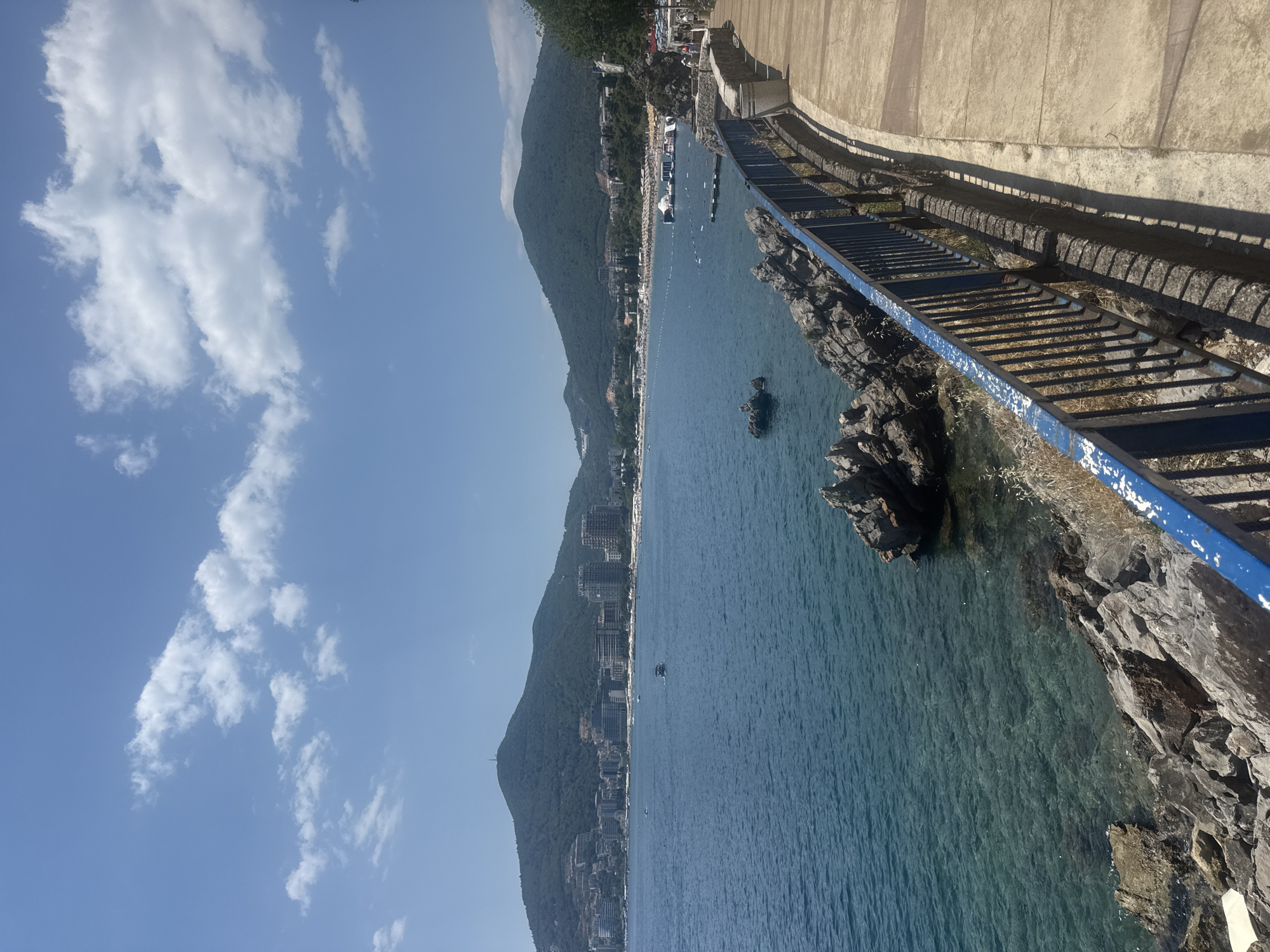 The image depicts a scenic view of the coastline in Budva, Montenegro. In the foreground, a paved pathway curves along the edge of the sea, bordered by a weathered blue metal railing. Rocky outcrops are visible at the water's edge, with the clear turquoise water revealing the seabed.
The middle ground features the blue sea with a few small boats. Beyond the water, a long, crescent-shaped beach is lined with numerous beach chairs and umbrellas, indicating a populated recreational area. Various buildings, including hotels and apartments, are situated along the hillside behind the beach, stretching towards the two prominent, green, tree-covered mountains that dominate the background. The sky is clear blue with scattered white cumulus clouds.