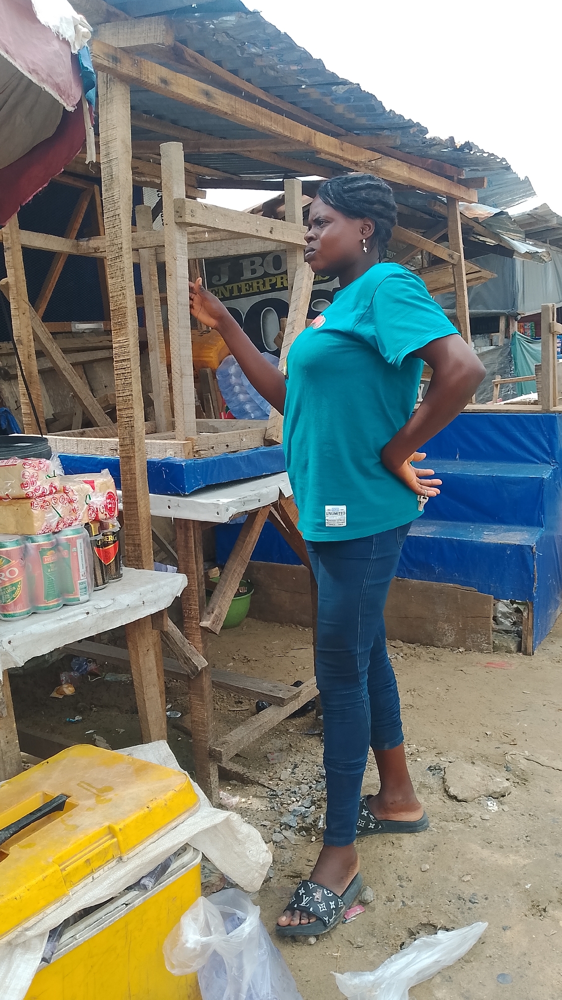 A Black woman stands in front of a makeshift market stall in Warri, Nigeria. She is wearing a teal t-shirt and blue jeans, with black slides adorned with a white monogram pattern. Her left hand is on her hip, and her right hand is extended, appearing to gesture. The stall is constructed of weathered wooden planks and has a corrugated metal roof. To the left of the woman, on a lower shelf, are several cans of what appear to be alcoholic beverages, and stacks of wrapped goods. Behind her, a blue tarp covers raised platforms, and other market structures are visible in the background. The ground is dirt and littered with debris.