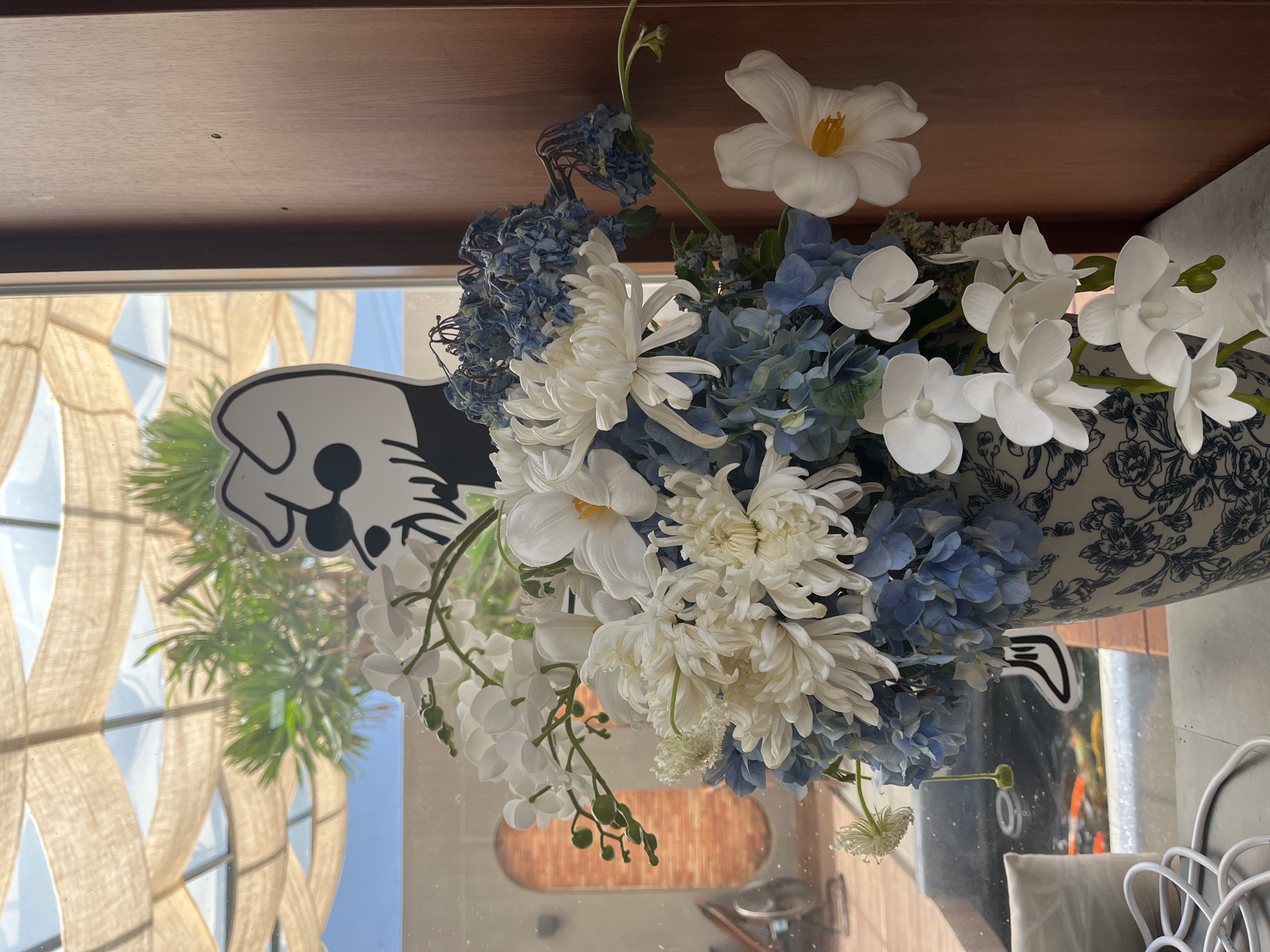 A still life composition features a vibrant arrangement of artificial flowers set on a reflective surface, likely a glass table or windowsill, in an indoor setting in Đà Nẵng, Vietnam. The bouquet predominantly consists of white chrysanthemums, delicate white phalaenopsis orchids, large white flowers with yellow centers, and clusters of blue hydrangeas, alongside some dried-looking blue florets. These flowers are housed in a decorative vase with a white background and intricate blue floral patterns.
Behind the floral arrangement, a large window offers a view of a bright, sunny day with a clear blue sky. Outside the window, a distinctive building facade or shade structure with a large, light-colored, open-weave or criss-cross pattern is visible, possibly made of wood or natural fibers. Green foliage is also seen through the window, indicating outdoor plant life.
Adding a whimsical touch to the scene is a black and white cutout or sticker in the shape of a dog's head, positioned in front of the window. The reflective surface below the flowers captures reflections of both the outside world and elements of the interior space, including warm, wooden-toned structures, possibly part of a building's architecture or furniture, and an arched feature in the background. The bright, natural lighting suggests it is daytime, likely morning or afternoon, in a serene and well-lit environment. There is no visible text.