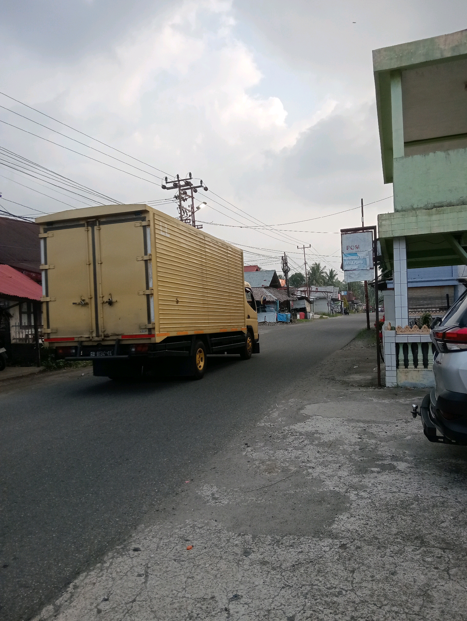 The image captures a street scene in Campago, Indonesia. A large, yellow box truck is driving on the right side of a paved road. The truck's rear doors are closed and secured with latches. The truck has a license plate that reads "BA 8134 GX". To the left of the road, there are some houses with red roofs and other buildings. A utility pole with multiple wires runs overhead. On the right side of the road, a light green building with a covered porch is visible, and a silver SUV is parked in front of it. A sign is mounted on a pole in front of the building. The sky is overcast with clouds. The road surface shows signs of wear and tear. The overall environment appears to be a rural or suburban area.