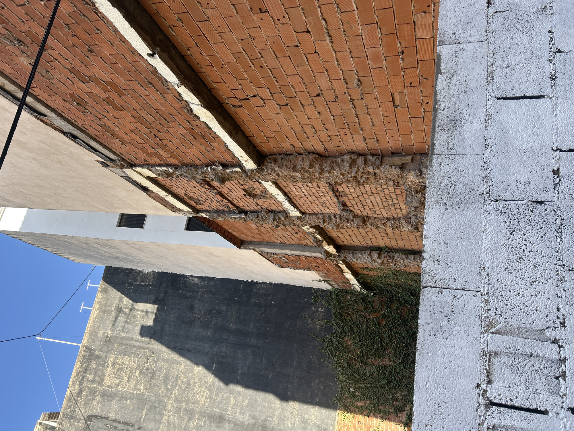 This image captures an outdoor scene with a focus on architectural elements and construction. The foreground features a wall constructed from large, white, textured blocks, with visible lines indicating individual units. Adjacent to this is a section of exposed red brickwork, which appears to be part of a wall or ceiling. The bricks are laid in a traditional pattern, and some of the mortar is crumbling, suggesting ongoing construction or disrepair.
The background reveals more of the surrounding environment. A light-colored building with windows is visible on the left, partially obscured by a dark, textured wall. This dark wall has a rough surface and shows signs of weathering. Sunlight casts a strong shadow across this wall, indicating it is likely daytime. Further to the left, a clear blue sky is visible, along with electrical wires and two white vertical structures that resemble antenna mounts.
A significant portion of the image is dominated by the red brickwork, which is angled upwards, giving a sense of looking up or into a recessed space. There are patches of plant growth, specifically a dense vine, clinging to the lower sections of the brick wall on the right side. The overall impression is one of an urban or semi-urban setting undergoing some form of development or renovation. The lighting suggests a sunny day, possibly late morning or early afternoon, given the distinct shadows. No people or explicit text are visible in the image. The location context provided is Castelló De La Plana, Spain.