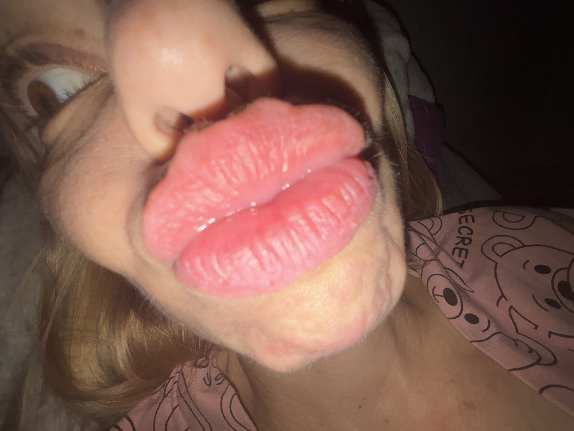The image is a close-up, low-angle shot of a person's face, focusing on their mouth and nose. The person's lips are parted, revealing a tongue. The lighting is dim, creating shadows and highlighting the textures of the skin and lips. The person appears to be wearing a pink garment with a pattern of bears and the word "SECRET" visible. The background is dark and out of focus, suggesting an indoor setting. The scene does not contain any specific landmarks or elements that would allow for the identification of a location within the city of Sinyavino, Russia.