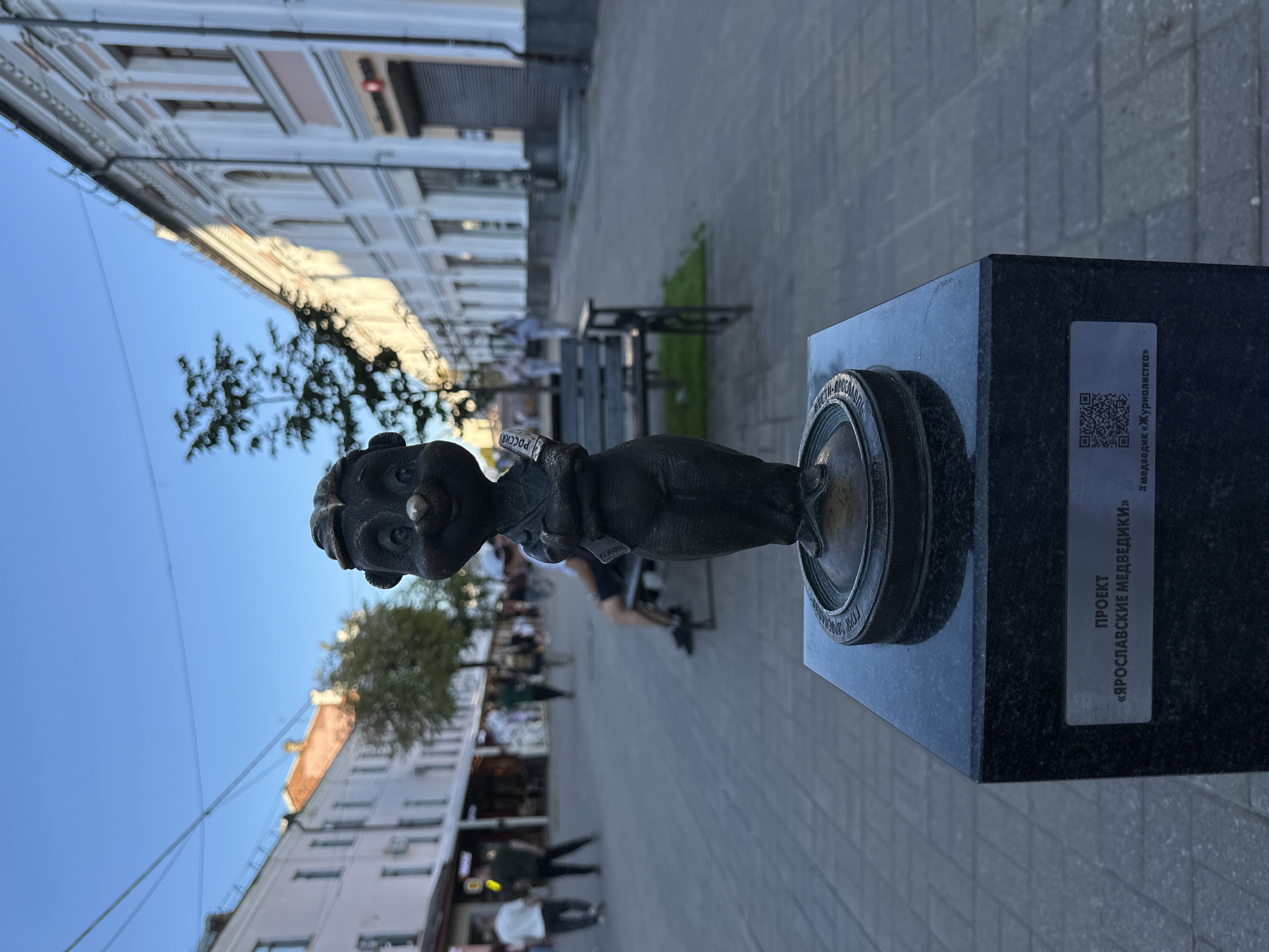 A bronze statue of a cartoonish bear wearing a vest and holding a small object is positioned on a black granite pedestal in the foreground. The statue is located outdoors on a paved walkway lined with buildings. The bear is depicted with its arms crossed and a slight smile. On the pedestal, a metal plaque reads "ПРОЕКТ «ЯРОСЛАВСКИЕ МЕДВЕДИКИ»" and "#медведик «Журналистка»". A QR code is also visible on the plaque. In the background, people are walking along the street, and there are benches and trees. The buildings lining the street appear to be shops or businesses. The sky is clear and blue.