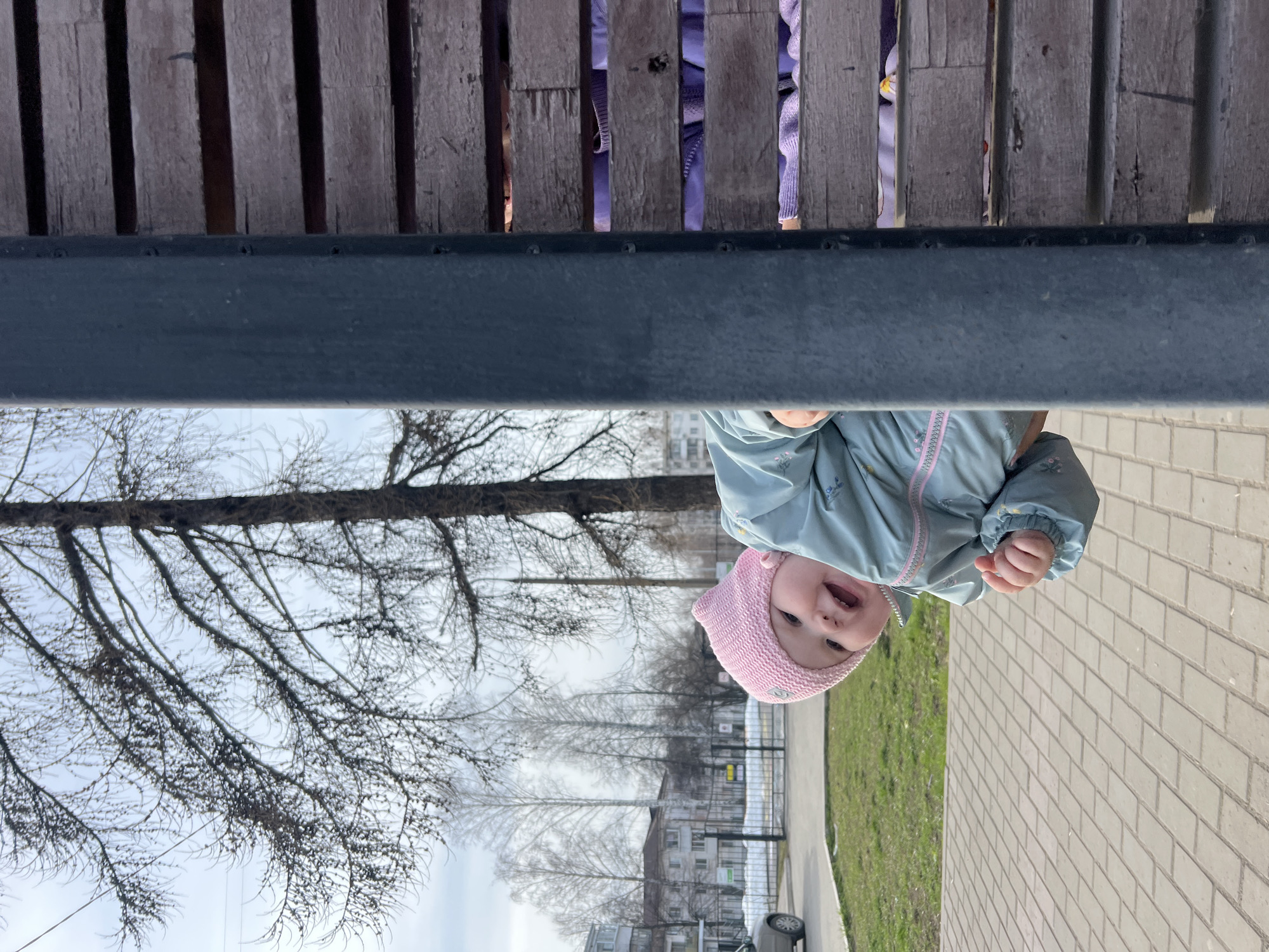 A baby wearing a pink hat and a light blue jacket is looking at the camera from under a wooden bench. The bench is located in a park in Chaikovsky, Russia, based on the surrounding architecture and the Russian language signage that is faintly visible in the background. The ground is paved with concrete blocks. A row of bare trees stands between the park and the buildings. The sky is overcast.