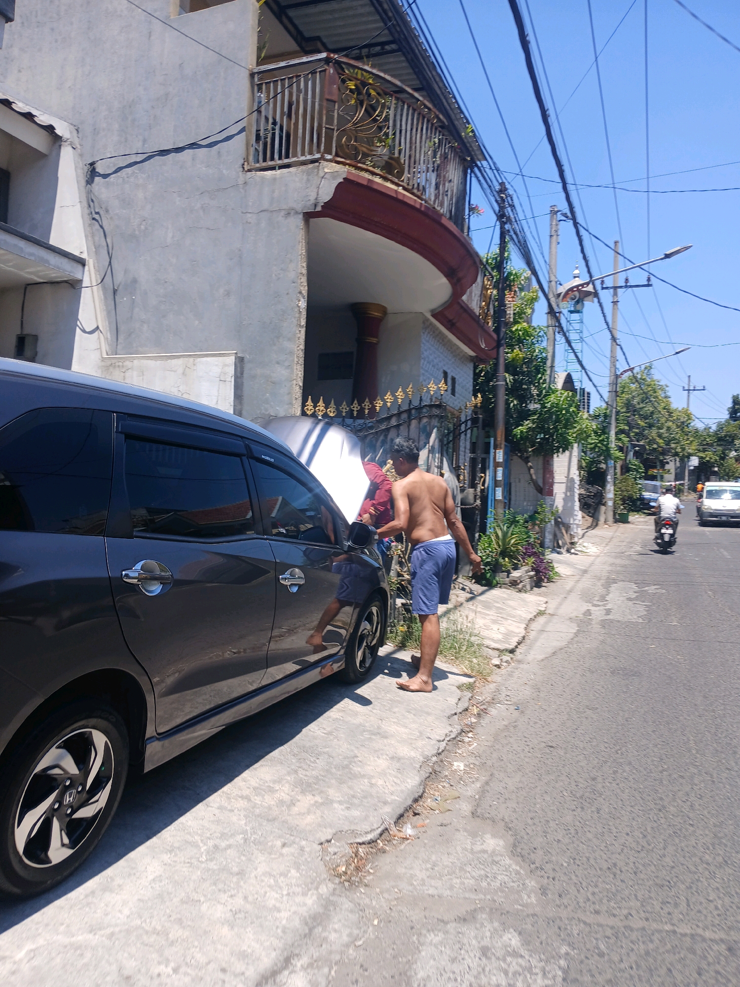 An outdoor, eye-level, medium shot captures a man standing next to a dark grey minivan on a sunny day. The minivan is parked on a concrete sidewalk next to a paved road. The hood of the minivan is open, and the man, wearing blue shorts and no shirt, appears to be working on the engine. He is facing away from the camera and towards the open hood. In the background, a multi-story building with a balcony is visible, along with trees and power lines. The sky is clear and blue. The street is visible on the right side of the image, with a white car and a motorcycle in the distance. The scene is set in Surabaya, Indonesia, indicated by the style of the building and the presence of a mosque-like structure in the background.