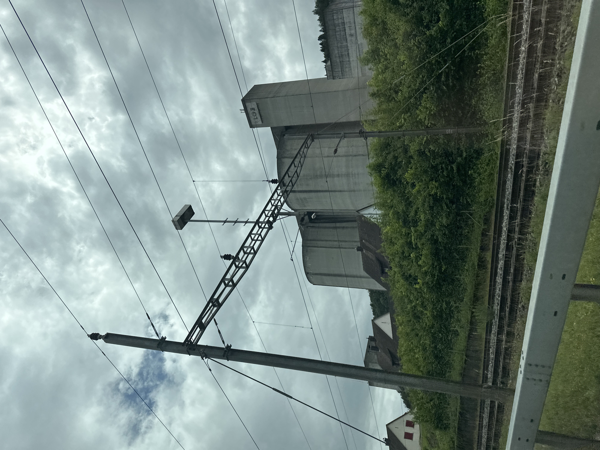 The image, oriented upright, captures an overcast daytime scene alongside a railway line in Zurzach, Switzerland. In the foreground and midground, a substantial concrete pole supports an overhead railway electrification system (catenary), featuring multiple wires and a metal lattice structure. A rectangular light fixture is mounted on the pole.
To the right of the pole, partially concealed by dense green trees and bushes, stand two prominent grey concrete structures. The taller one is a rectangular, tower-like building with faded black graffiti on its upper facade, appearing to spell "ROTA". Below and to its right is a second, shorter and more rounded concrete building, likely an industrial or agricultural silo.
Further right, multiple railway tracks run parallel, visible through and behind a metallic barrier, alongside grassy areas and more green foliage. In the lower right background, parts of houses with distinctive red-tiled roofs can be seen nestled among the trees. The sky is entirely covered with thick, grey clouds, indicating an overcast day. No people or specific activities are visible, presenting a static snapshot of the railway infrastructure and surrounding landscape.