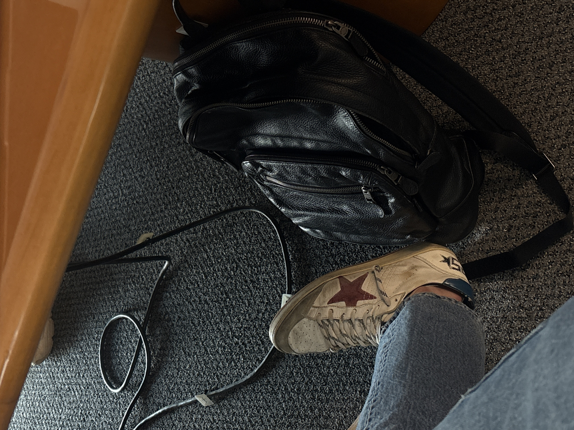 The image depicts an indoor scene on a gray carpeted floor. A black leather backpack sits next to a person's leg clad in denim jeans and a white sneaker with a red star. A black cable, secured by white cable clips, runs along the carpeted floor. The scene includes part of a wooden desk or table. The location cannot be determined from the information provided.