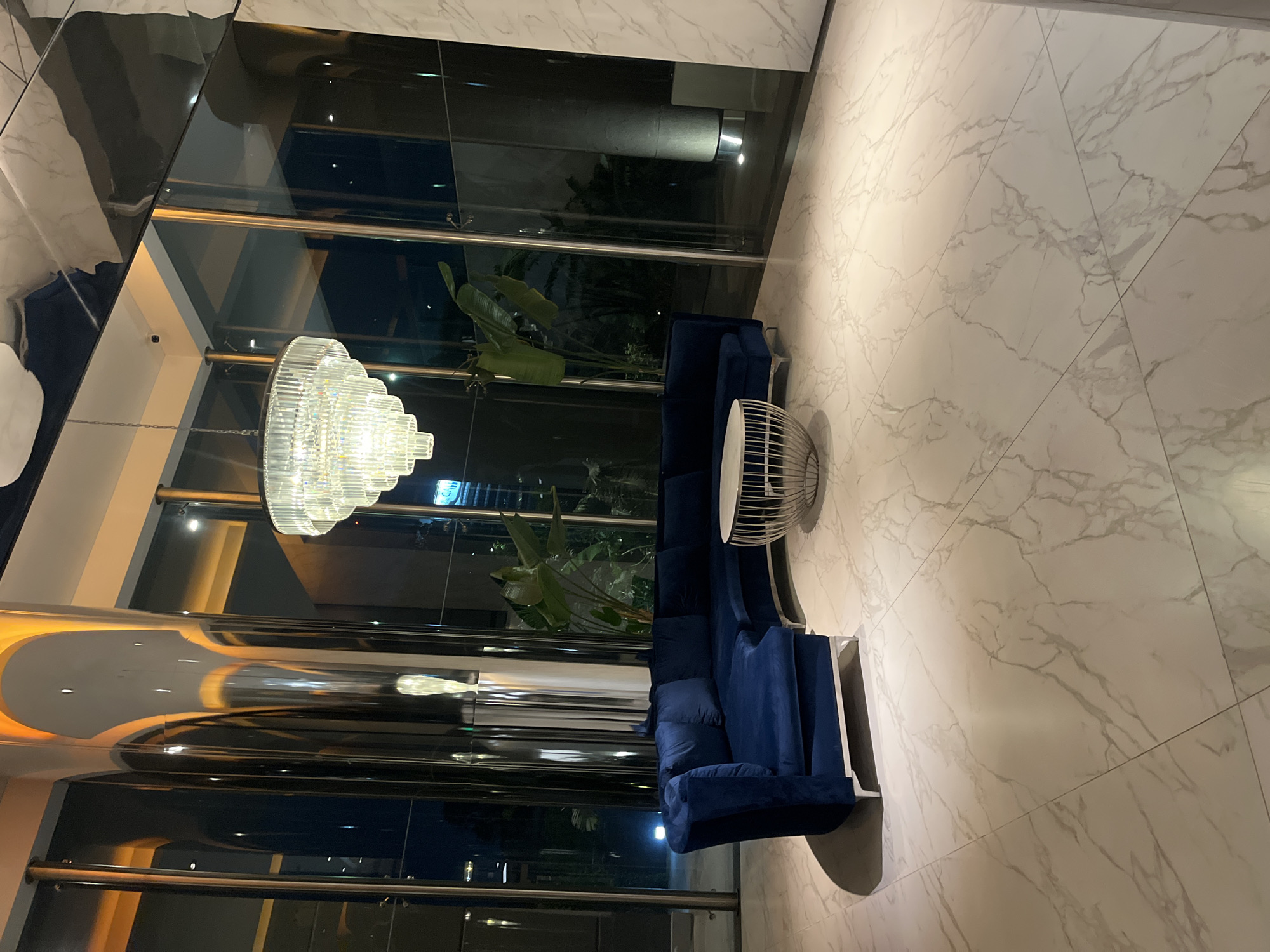 The image captures an interior space, likely a lobby or reception area, characterized by modern design. A prominent feature is a multi-tiered crystal chandelier that illuminates the scene. The floor is covered with large-format white marble tiles with grey veining. A blue velvet sofa with white frame accents is positioned adjacent to a small, cylindrical metal side table. Large windows offer a glimpse of greenery, possibly indoor plants or an exterior garden. Reflective surfaces are apparent, contributing to the overall contemporary aesthetic.
There is insufficient information in the image to identify the location in Buenos Aires, Argentina.