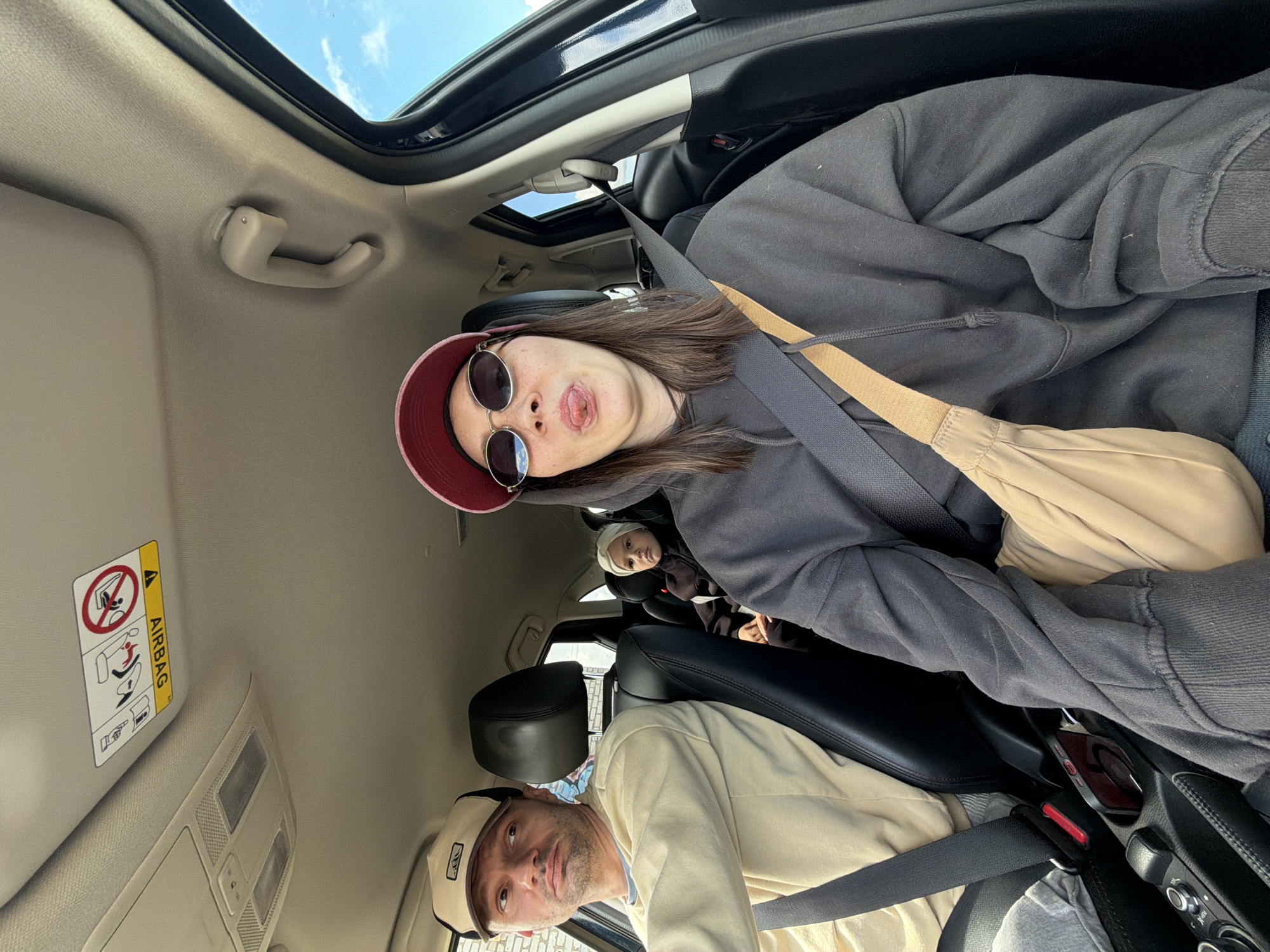 The interior of a car is visible in the image, with four people inside. A woman in a red visor and round sunglasses is in the front passenger seat sticking her tongue out. She is wearing a dark gray hoodie and a tan shoulder bag. The driver is wearing a light tan shirt and a baseball cap. In the back, there is a child in a white hat and another person in a dark outfit. Everyone is wearing seatbelts. A sunroof is visible above the front seats, and an airbag warning label is on the sun visor. The location of the image is unknown.