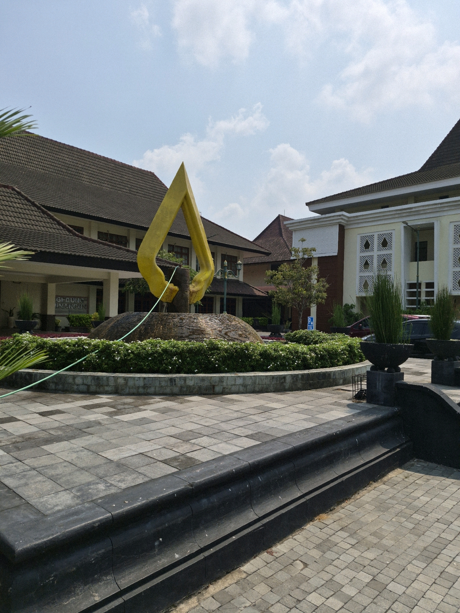 The image shows a monument with a golden sculpture in the foreground, located in front of a building complex. The sculpture is abstract and appears to be made of polished metal. It is situated on a circular base covered with decorative stones and surrounded by a manicured garden with small white flowers and green bushes. A light green hose or cable runs across the garden and the paved area.
Behind the monument, there are several buildings with tiled roofs and beige-colored walls. One building has an entrance with a sign that reads "GEDUNG KEMUNING". Another building features decorative square windows. The architectural style suggests a public or institutional building, possibly a government office or educational facility.
The ground in front of the buildings is paved with square gray tiles. A dark-colored raised platform with curved edges is in the immediate foreground, leading up to the tiled area.
The environment appears to be outdoors on a sunny day, with a clear blue sky and scattered white clouds. There are some plants in large black pots on the right side of the image, and a few cars are visible in the background, parked near the buildings. There are no people in the frame.
The location is the front area of the Wates City Hall in Wates, Kulon Progo Regency, Yogyakarta Special Region, Indonesia.