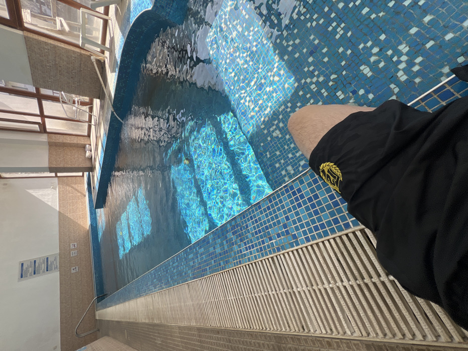 The image depicts an indoor swimming pool facility located in Banite, Bulgaria, viewed from a first-person perspective. The lower torso and bare legs of an individual wearing black swim shorts are visible in the foreground, seated at the edge of the pool. The shorts feature a yellow embroidered emblem, possibly a stylized "E.T." within a laurel wreath, on the left thigh.
The pool is an irregularly shaped body of water with various depths, lined entirely with small blue mosaic tiles. Multiple submerged steps are visible within the pool. Clear blue water fills the pool, reflecting bright sunlight that streams in from large windows positioned along the right wall. A metal handrail is affixed to the pool's edge at the far left. A water feature, possibly a spout or small waterfall, feeds into a lower, distinct section of the pool on the right.
The immediate pool surround features a light-colored, slatted drainage grate running parallel to the pool's edge. The walls of the facility are constructed with light brown rectangular tiles on the lower half and white painted surfaces on the upper half. Informational signs are mounted on the white wall in the upper left background. Two white plastic chairs are visible near the windows on the right side of the facility. The exterior environment, partially visible through the large windows, suggests other structures or buildings.