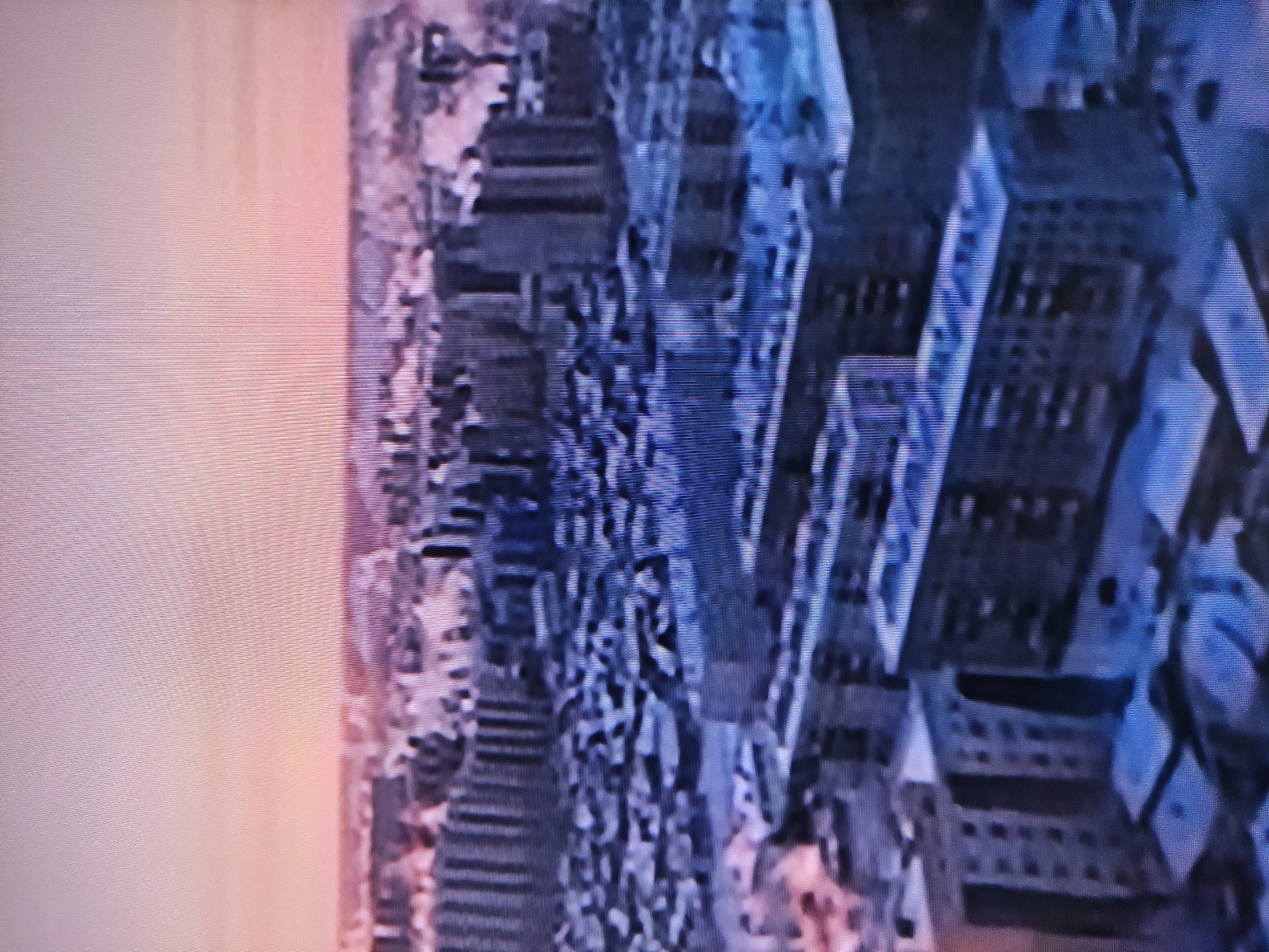 The image shows a cityscape under a sunrise sky. Tall buildings are visible, possibly residential or office structures. The city is likely Pavlodar, Kazakhstan, given the prompt. The buildings display repetitive window patterns. The sky fades from pale pink near the horizon to pale blue above the buildings. The image quality is grainy or pixelated.