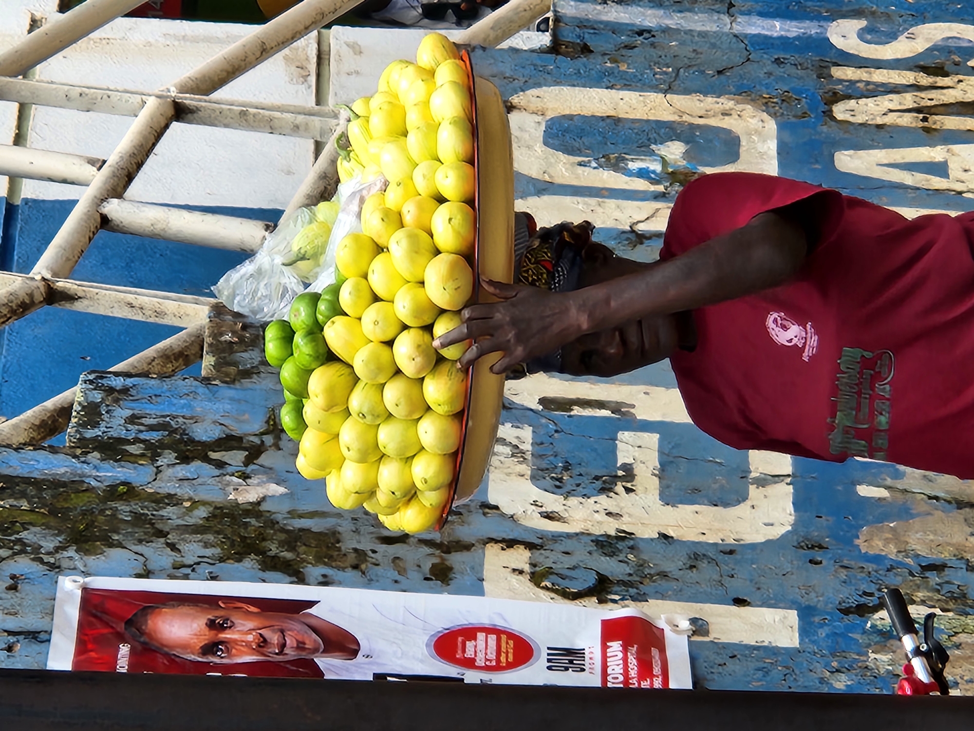 A woman is shown from the chest up, wearing a red t-shirt and a patterned headscarf, balancing a large tray of yellow fruit on her head. The fruit appears to be a mix of lemons and possibly some gourds. The woman's face is partially obscured by the tray. The background consists of a distressed, blue and white painted wall with peeling paint and visible cracks. To the left of the woman, a portion of a poster featuring a man's face is visible, with text in red and white. To the right, metal railings are present against the wall. A bicycle handlebar and part of a red brake lever are visible in the lower left corner. The lighting suggests it is daytime, with harsh shadows.
