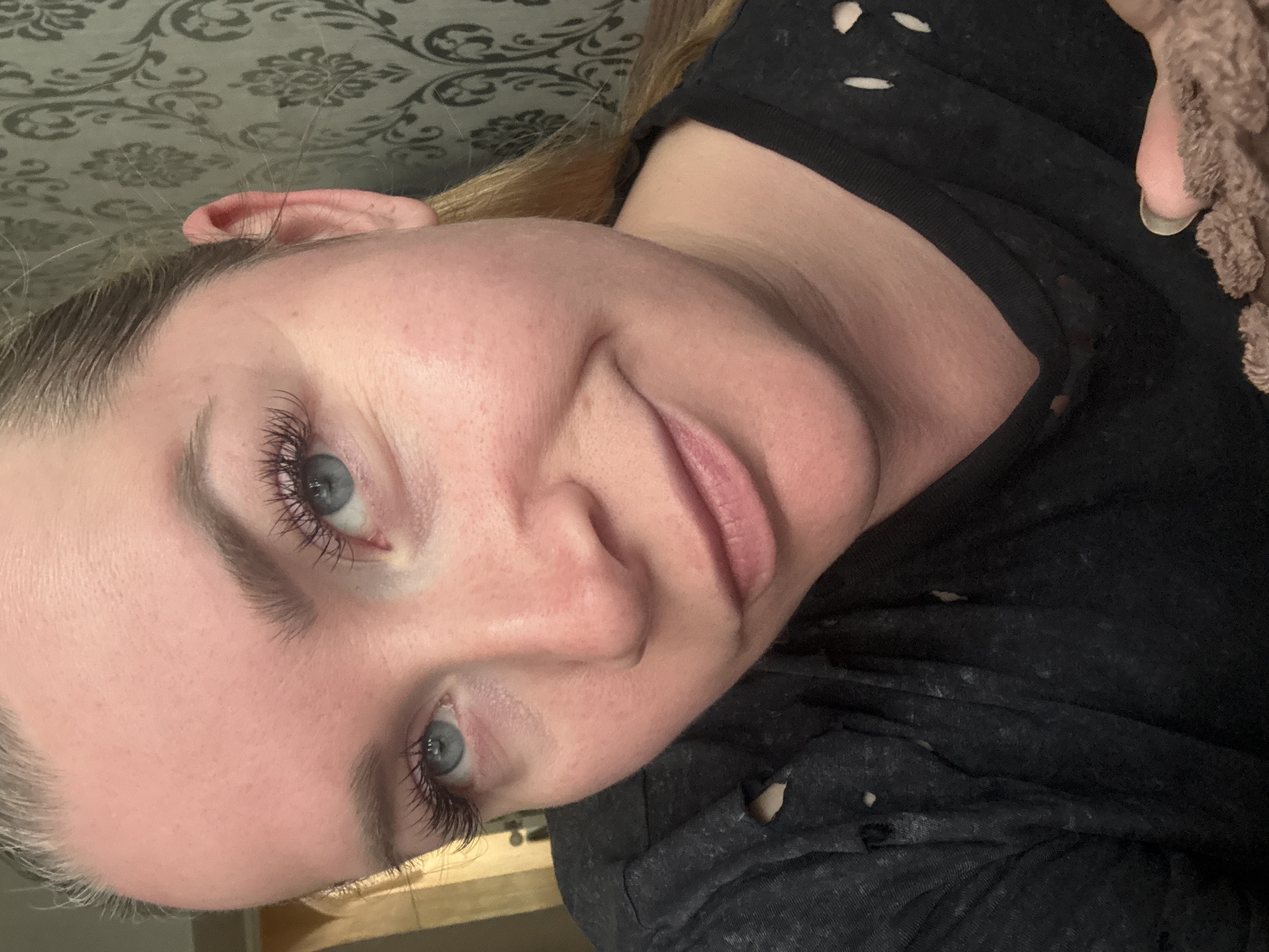A woman is angled in the frame from a low perspective. Her face is pale with light eyebrows and eyelashes. Her eyes are light blue. Her lips are pink. She wears a black shirt with multiple small holes. The background consists of a dark patterned surface. The image doesn't provide information to identify the location.