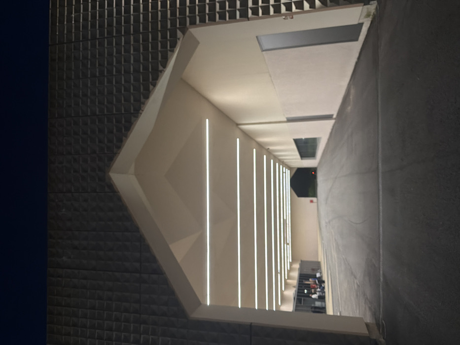 The image displays a modern architectural structure at night in Penafiel, Portugal. The central feature is a large, gabled archway, functioning as an illuminated covered passage or entrance. The building's exterior surfaces, above and flanking the archway, are clad in dark, possibly charcoal-grey, square-patterned relief tiles. The interior of the archway features light-toned, possibly off-white, faceted walls and ceiling. Nine parallel horizontal LED strip lights are embedded in the ceiling, providing consistent illumination along the passage.
On the left side of the archway, a glass-fronted section reveals a group of approximately four to five individuals, some seated, within what appears to be an interior waiting area. The ground beneath the archway consists of dark asphalt pavement, exhibiting subtle tire marks. A linear drain grate is visible across the pavement in the immediate foreground. The interior right wall of the passage is light-toned, featuring a long, horizontal window providing a view into an adjacent space, and further along, a dark grey vertical door. The passage extends into the distance, terminating in another dark, gabled opening, within which a single red light source is discernible.