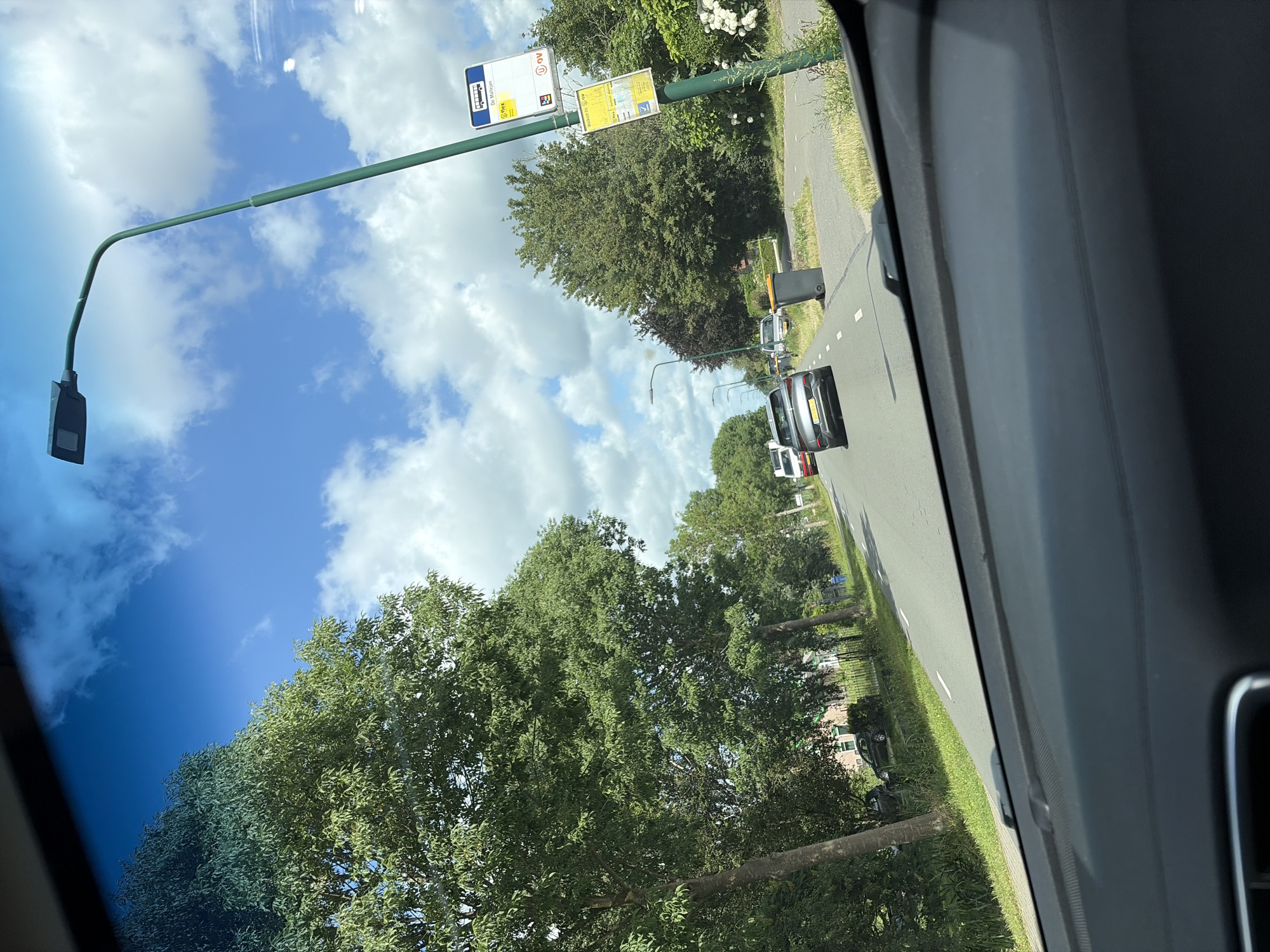 The image is taken from the perspective of someone inside a vehicle, looking out through the windshield. The scene depicts a sunny day in Maarssen, Netherlands, with a clear blue sky dotted with fluffy white clouds. A road stretches into the distance, lined with lush green trees on both sides.
In the foreground, the dashboard of the car is visible. On the right side of the road, a modern streetlight with a curved arm stands tall. Attached to the streetlight pole are two yellow signs, one at the top depicting a bus icon and text, and another below with more text and a bus icon. A grey garbage bin is situated on the grass verge next to the road, near the signs.
Further down the road, several cars are visible. A silver hatchback is in the immediate lane ahead, followed by a white van and other vehicles further in the distance. On the left side of the road, houses and fences are partially obscured by trees. The overall impression is a suburban or semi-rural environment with active road traffic.