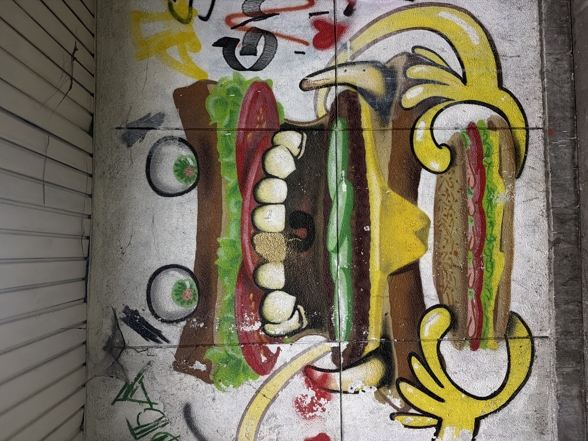This is a photograph of graffiti on a wall, likely in Sofia, Bulgaria. The main subject of the graffiti is a stylized sandwich or burger composed of various colors and shapes. The burger features layers that include brown bread, green lettuce, red tomato, white cheese or mayonnaise, brown meat, and yellow mustard. Additional elements of the graffiti include cartoonish eyes, abstract shapes, and lines in yellow, black, and red. The wall itself is textured and shows signs of wear and aging, with visible cracks and discoloration. The overall impression is of urban street art. The graffiti is applied to a wall with some adjacent white corrugated paneling. The graffiti spans across several concrete blocks that make up the wall.