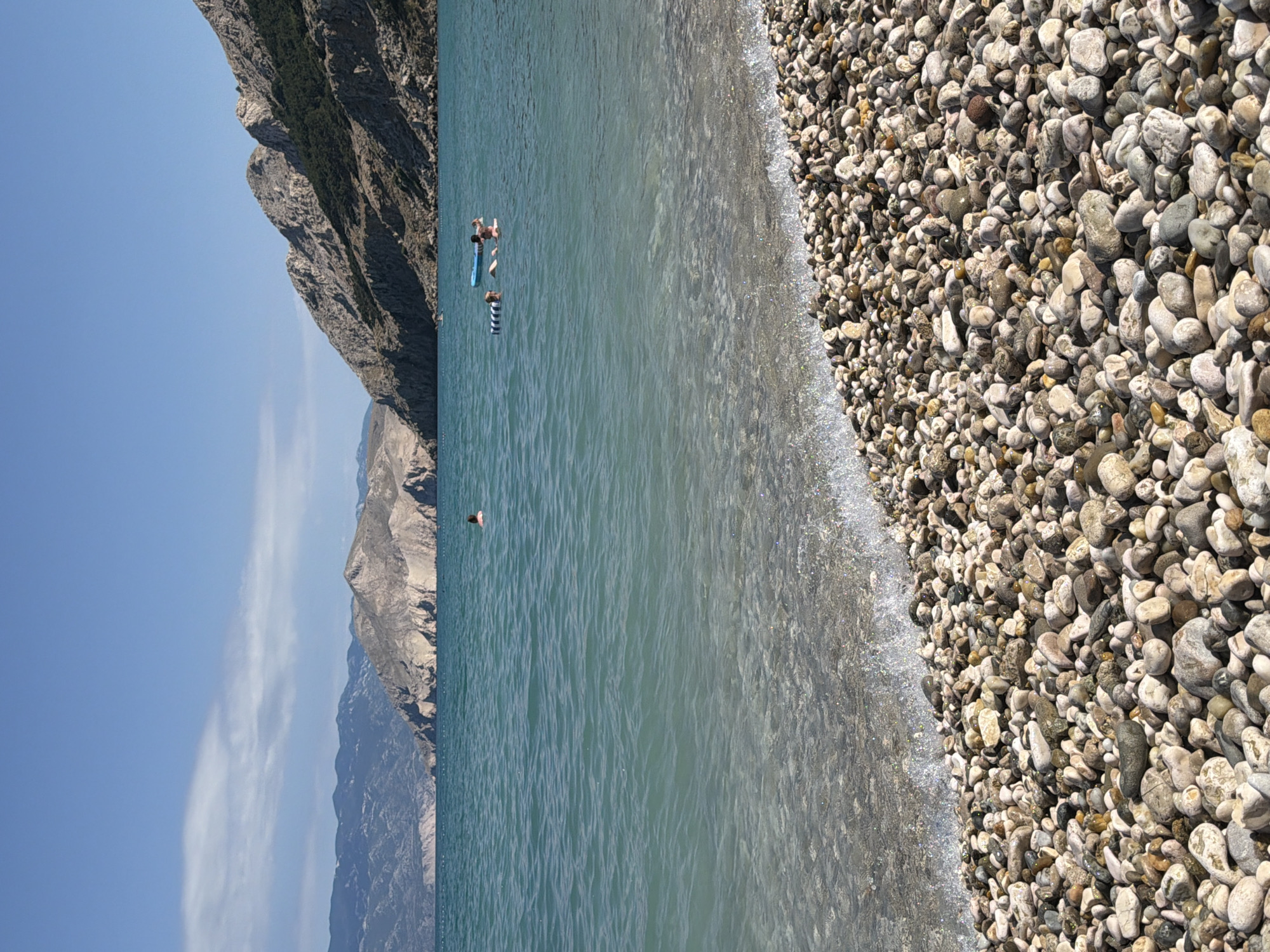 The image depicts a sunny day at a pebble beach in Općina Baška, Croatia. The foreground is dominated by a dense collection of smooth, multi-colored pebbles, with a gentle wave lapping at the shore, creating a shimmering effect of light. The sea is a clear turquoise, becoming deeper blue further out, with faint ripples on its surface. In the middle ground, several people are visible in the water. One person is on an inflatable raft, while another is swimming nearby. In the background, a rugged, mountainous coastline rises dramatically from the sea, characterized by rocky slopes and patches of sparse vegetation. The sky is a clear, vibrant blue with a few wispy white clouds.