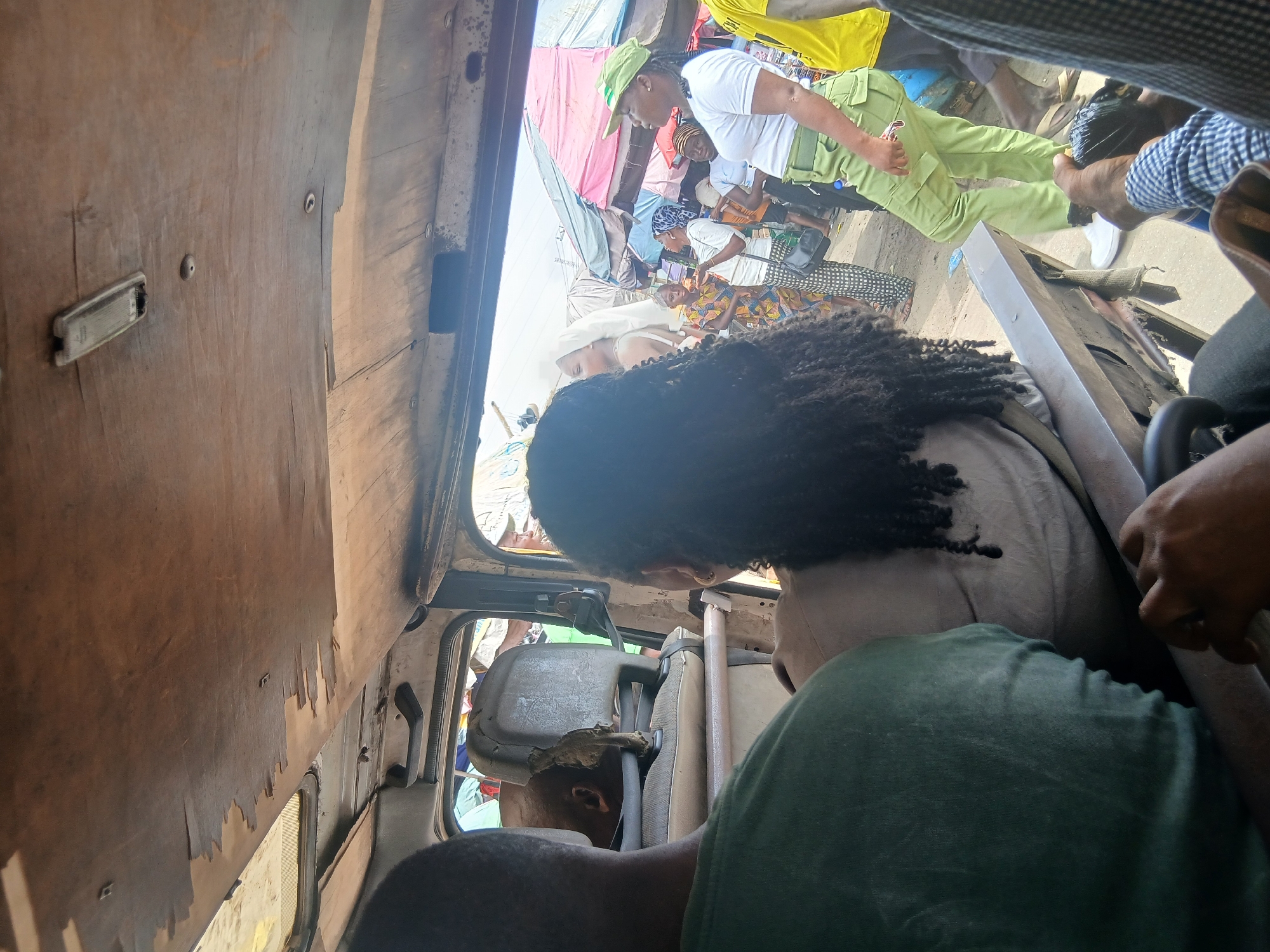 The image shows the interior of a vehicle, possibly a bus or a van, looking out towards a bustling market scene in Agani, Nigeria. The ceiling of the vehicle is made of worn, light-colored wood, with some sections peeling. A light fixture is visible on the ceiling. In the foreground, the backs of two people are visible. One person on the left wears a green shirt, and another person on the right has long, dark, curly hair and wears a grey shirt.
Through the open windows, the market is visible. People are walking and standing amidst market stalls covered by various colored tarps and umbrellas, including pink and white ones. A young woman wearing a green cap, white t-shirt, and green cargo pants, resembling a uniform, is prominently featured. She is looking to the right. Another person in the background wears a white headscarf and a patterned dress. A person in a yellow shirt is also visible. The ground appears to be paved, and there are indications of everyday market activity. The overall impression is that of a lively, public space in Nigeria.
