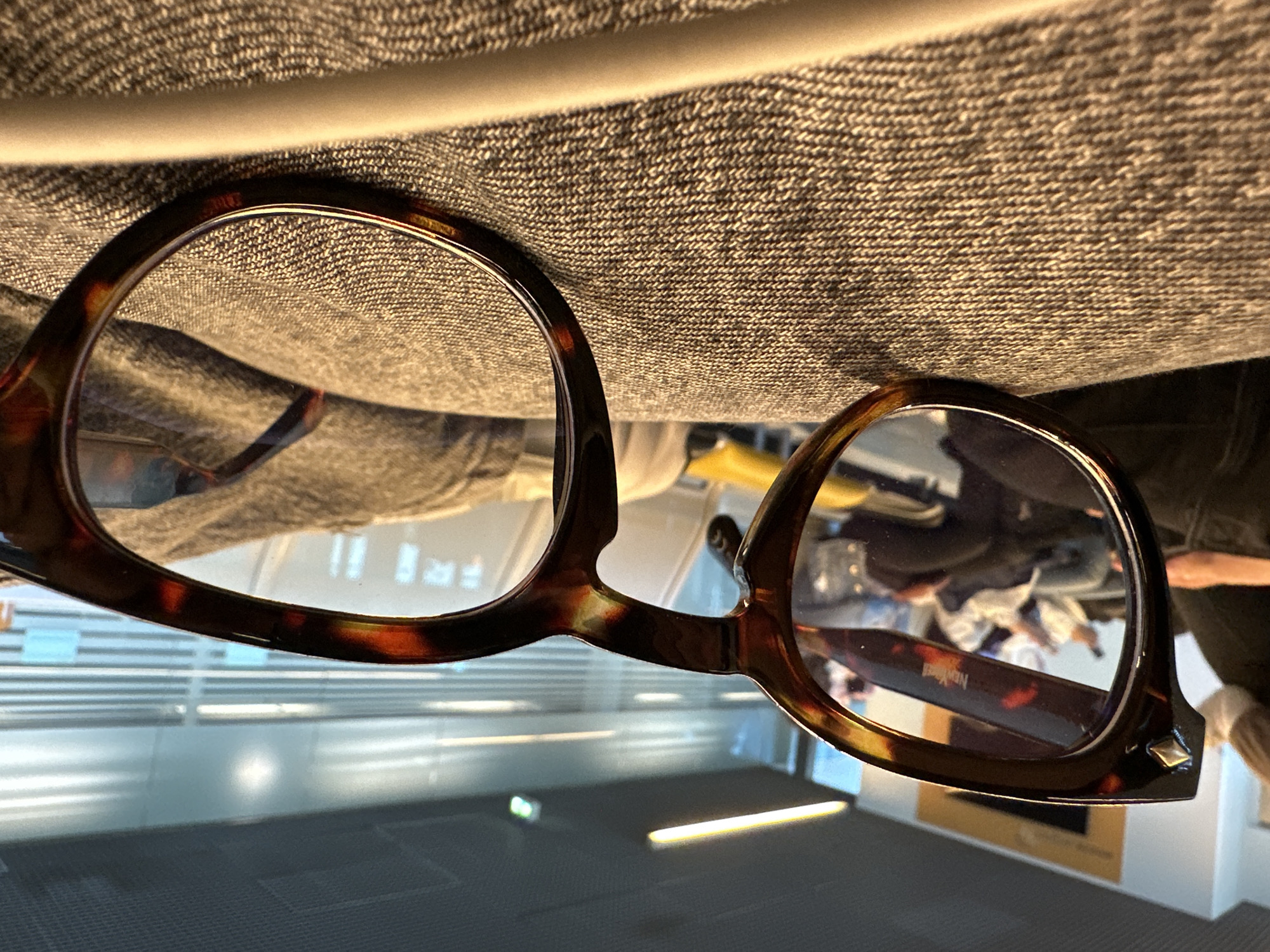 A pair of tortoise-shell patterned eyeglasses rests on a textured, grey fabric surface. The image is captured from a low perspective, looking through and past the frames into a bright, modern indoor environment.
Through the lenses and in the background, a light-colored, reflective floor shows a prominent horizontal streak of light. A light-toned railing or barrier stretches across the mid-ground. Beyond this, blurry reflections of several people are visible, suggesting a public area with activity. Distant, indistinct signage and an orange-brown object, possibly art or another sign, can also be perceived.
A notable detail on the eyeglasses is a small, metallic, pyramidal stud located on the outer corner of the right frame. The brand name "New York Montage" is clearly visible and legible on the inside of the right temple arm of the glasses. The contemporary architecture, polished surfaces, and reflections of individuals in the background, set within Weeze, Germany, indicate a bustling public concourse or waiting area, likely an airport terminal. The scene is well-lit, suggesting daytime.