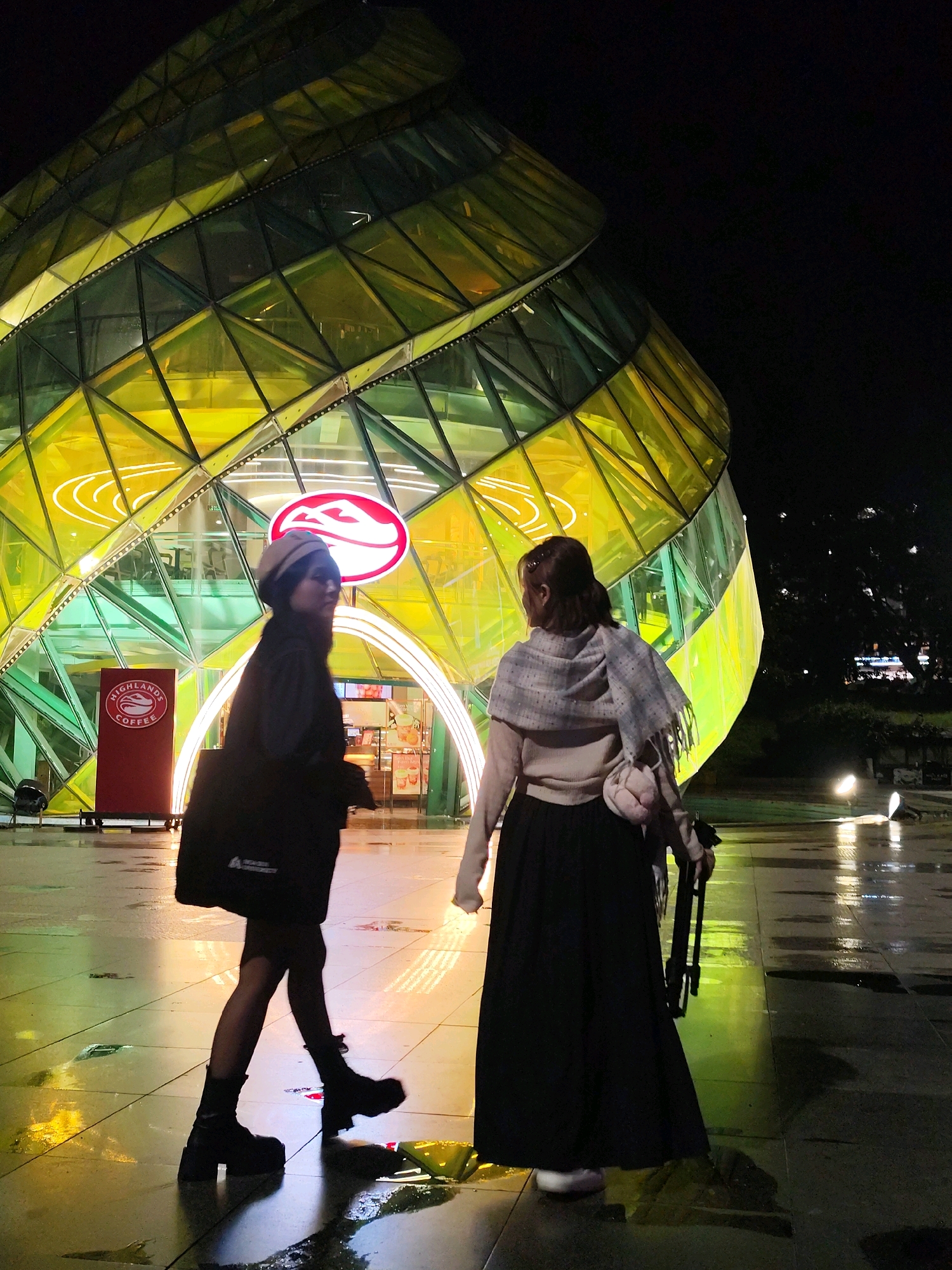 Two individuals are walking at night in front of a distinctive, iridescent, geometric building in Da Lat, Vietnam. The building, illuminated by internal yellow and green lights, has a unique, sculptural form composed of triangular glass panels. The ground in front of the building is wet and reflective, suggesting recent rain.
The person on the left, facing away from the viewer, is wearing a dark jacket, dark pants, and dark boots, with a light-colored beret. They are carrying a black tote bag. The person on the right, also facing away, is wearing a light-colored sweater, a dark long skirt, and a patterned scarf. They are carrying a small pink bag and what appears to be a foldable umbrella or cane. In the background, a red sign with a white circular logo, identified as "HIGHLANDS COFFEE," is visible on the building. The overall scene depicts an urban night environment with a focus on the illuminated architecture and the presence of pedestrians.